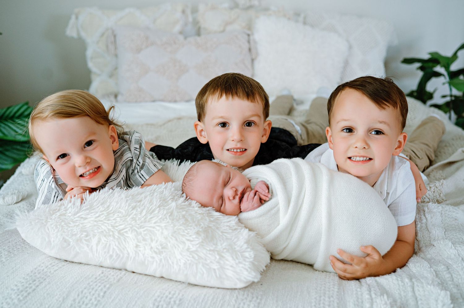 Children laying on white bedding smile together in a bright room with plants.