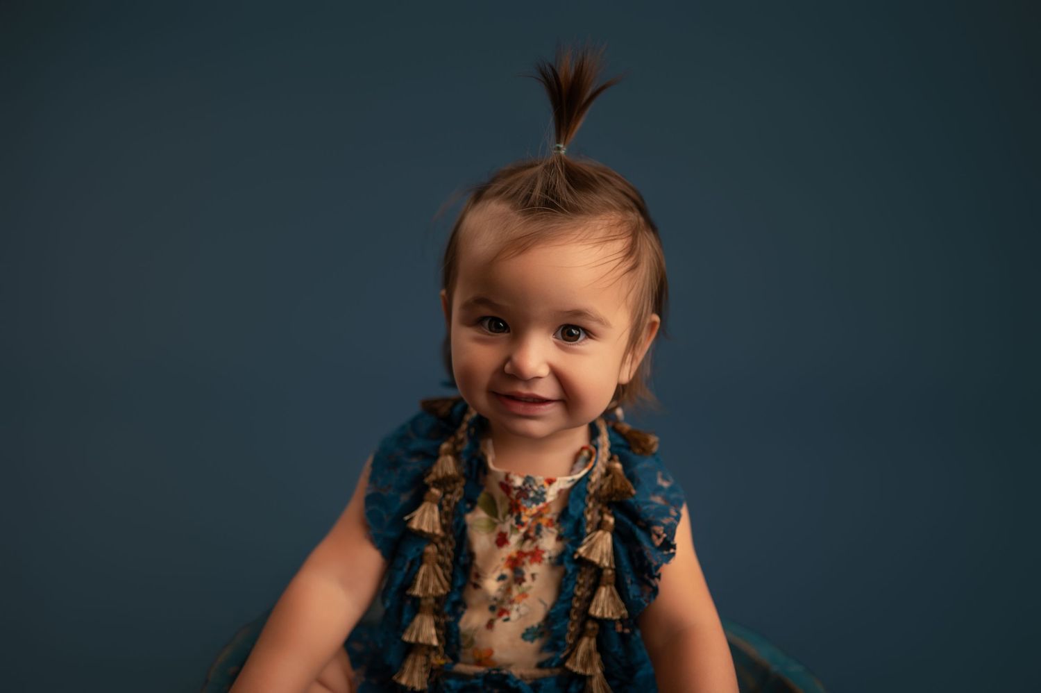 Smiling child wearing a navy blue dress with tassels and braided hair poses against dark teal backdrop.