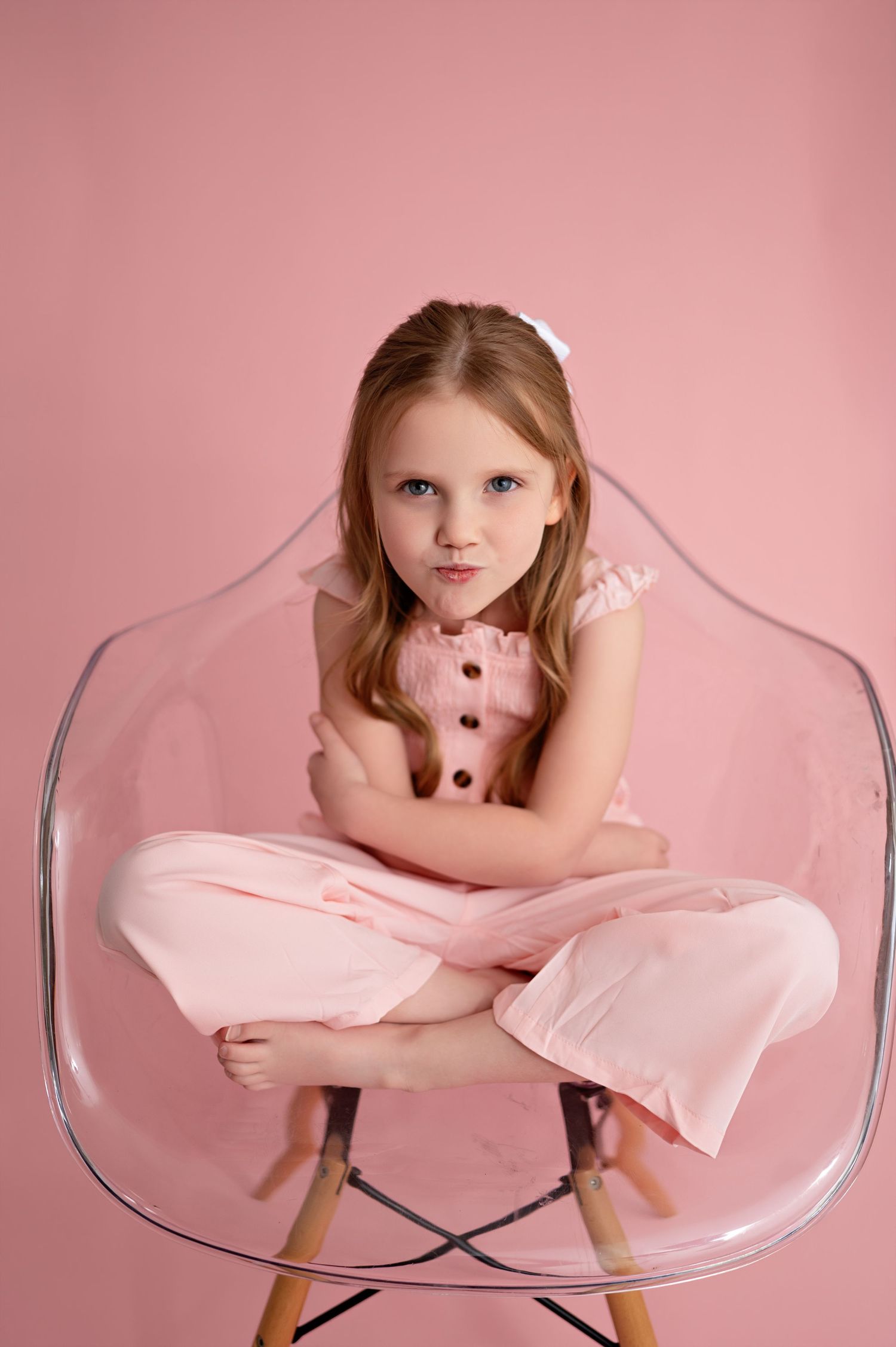 A person in a pink dress poses artistically on a clear acrylic chair against a pink background.
