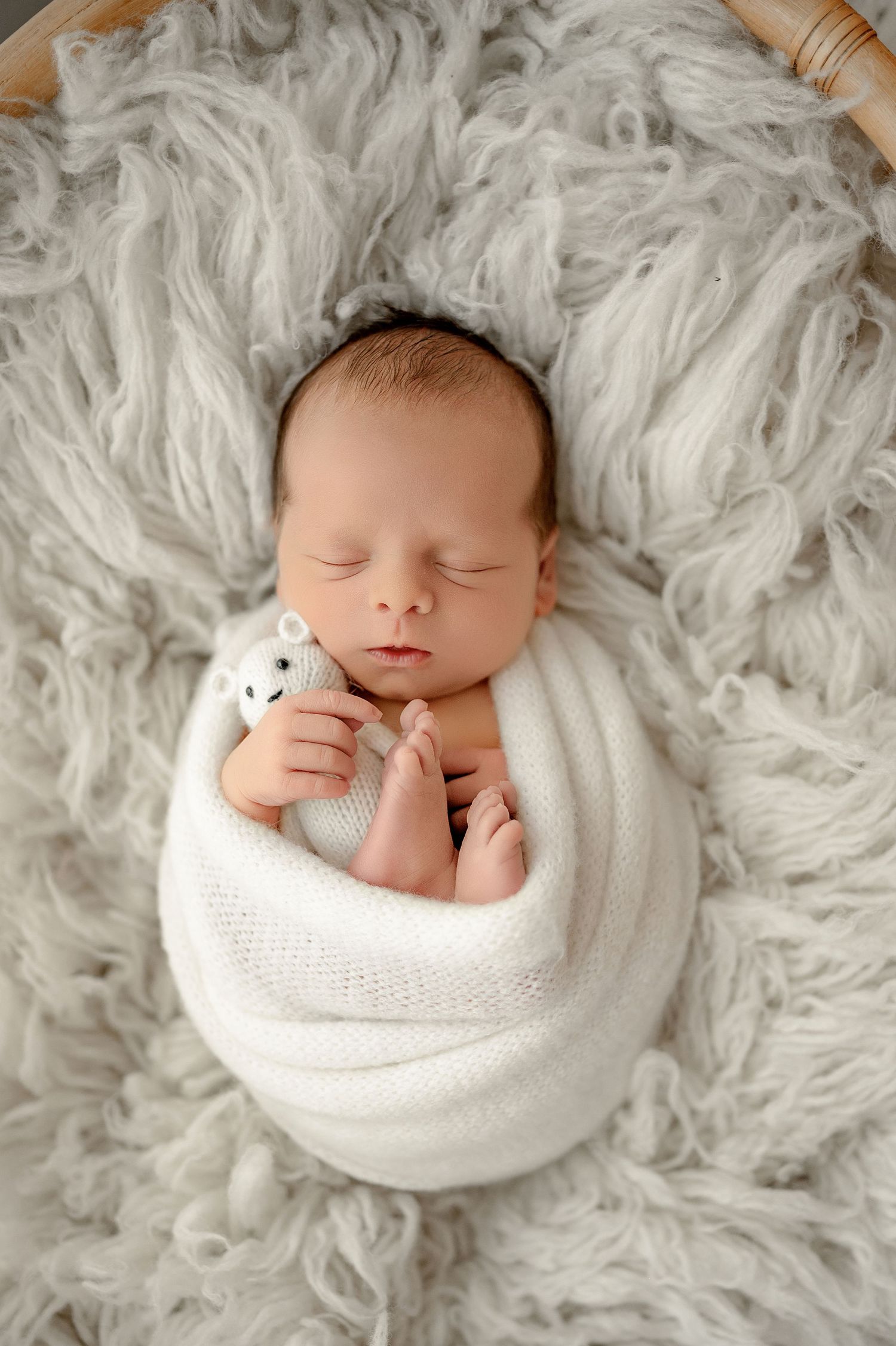 Sleeping newborn baby wrapped in soft white swaddle blanket lying on plush gray fur background.