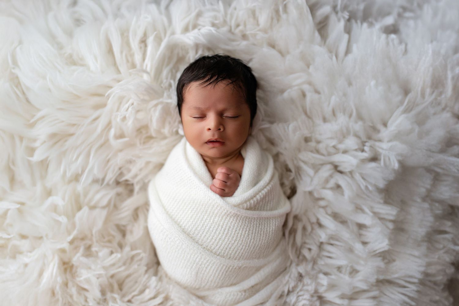 Peacefully sleeping newborn wrapped in white knit blanket lying on soft fluffy white fur backdrop.