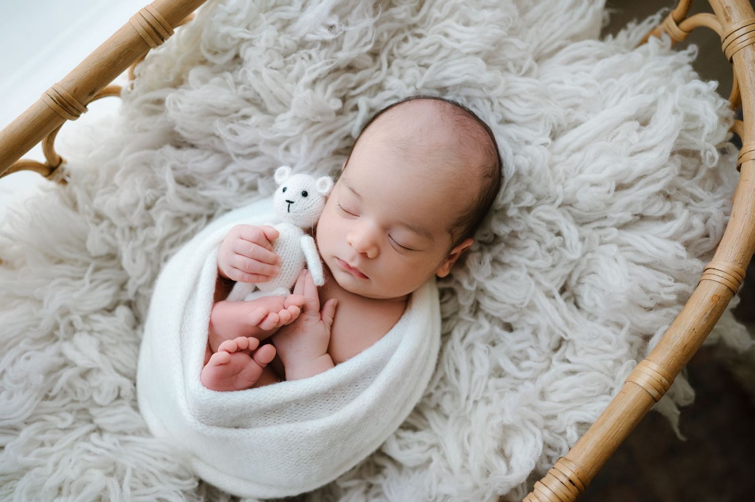 Newborn baby swaddled in white wrap sleeping in wicker basket with fluffy rug holding small teddy bear.