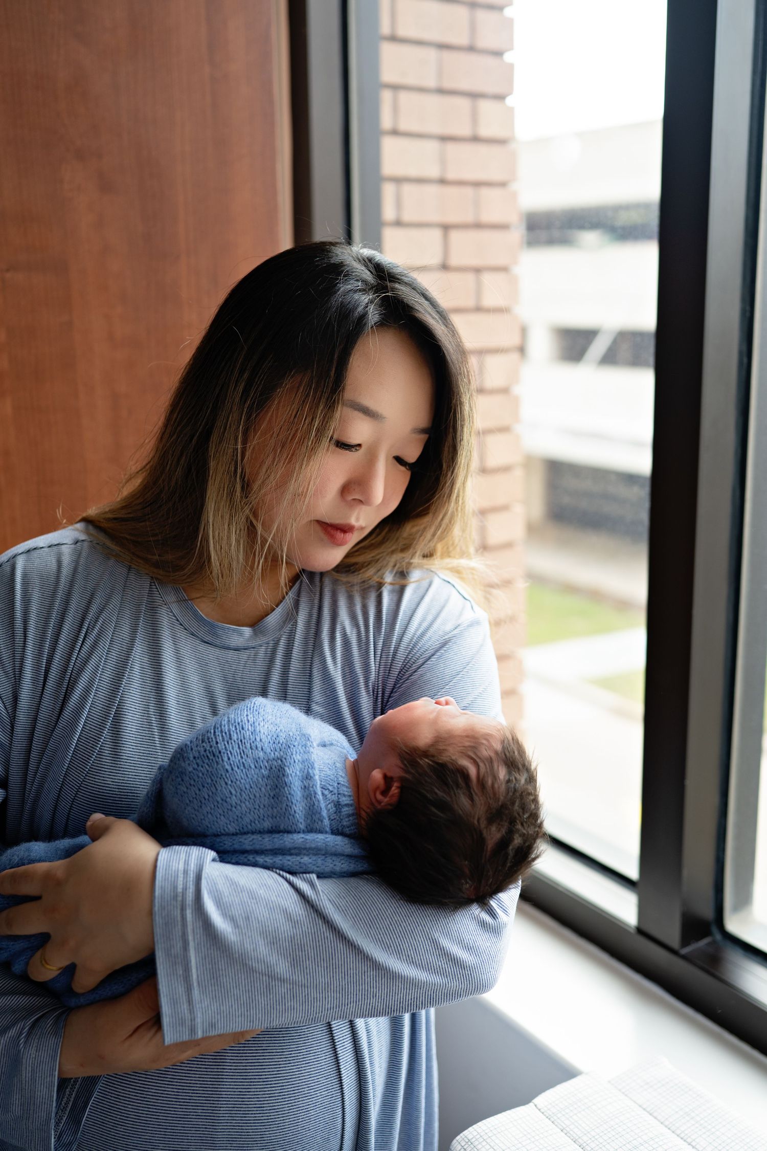 A tender moment by a window as a parent in gray clothing cradles a sleeping newborn baby in natural light.
