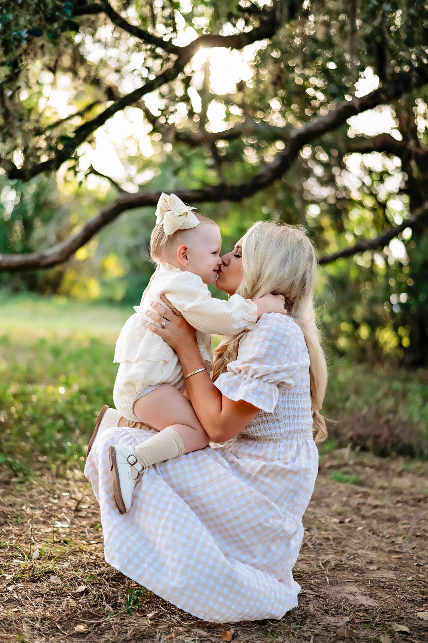 A tender outdoor portrait of parental love with a white gingham dress against a sunlit garden backdrop.