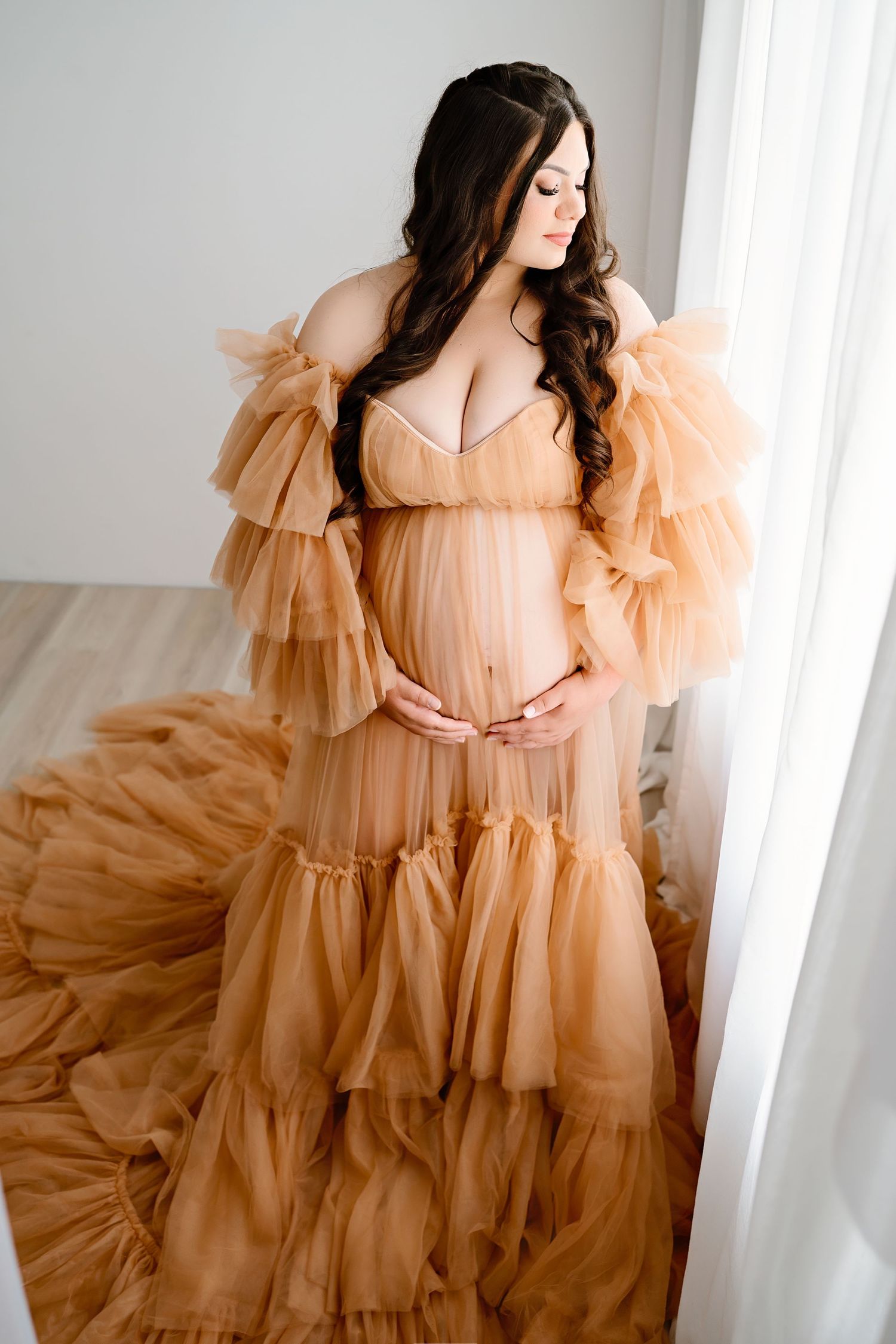 An elegant maternity portrait featuring a flowing peach-colored tulle gown in natural window light.