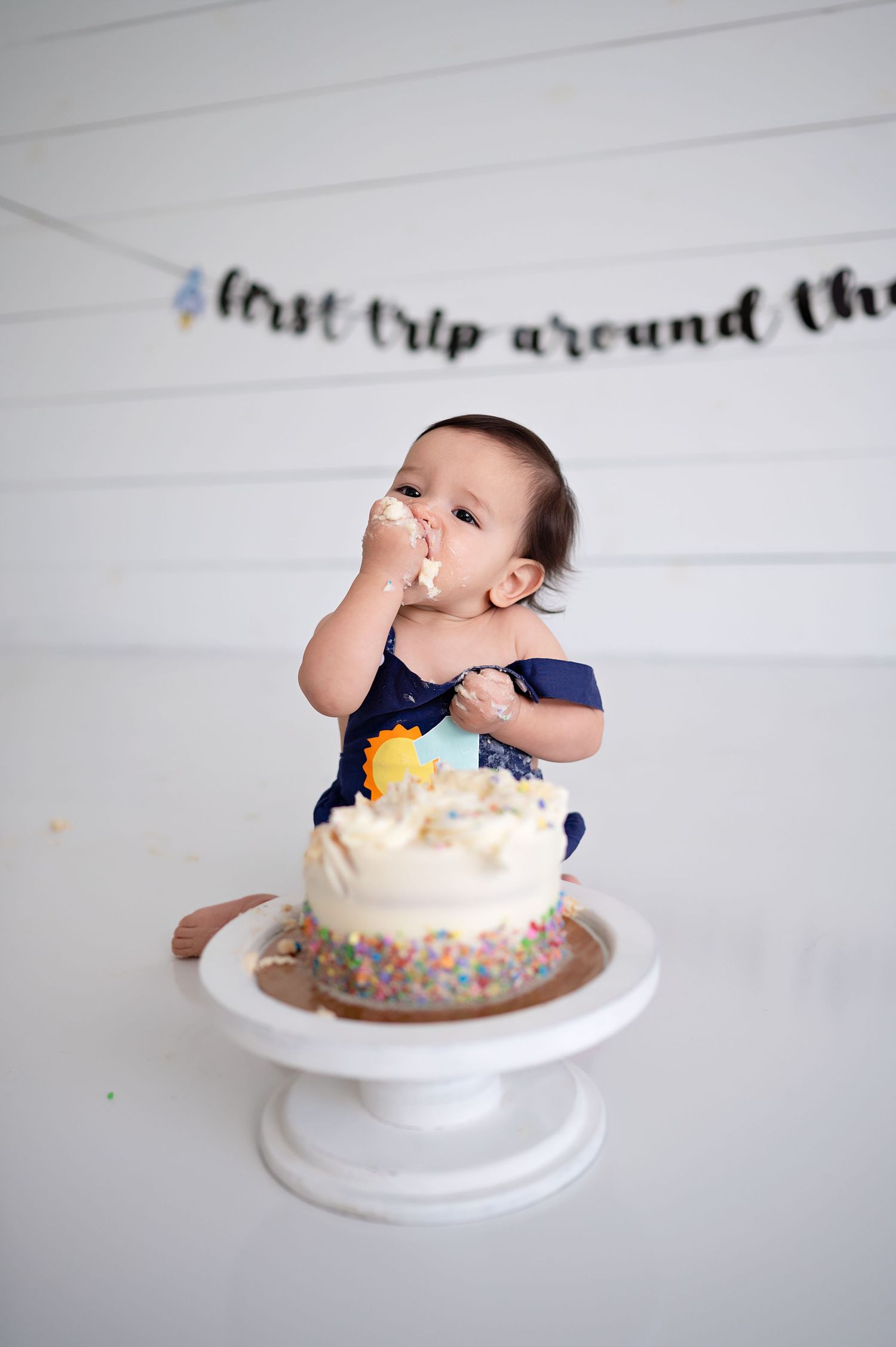 A baby in navy blue outfit sitting at a white cake stand eagerly eating and smashing into a birthday cake.