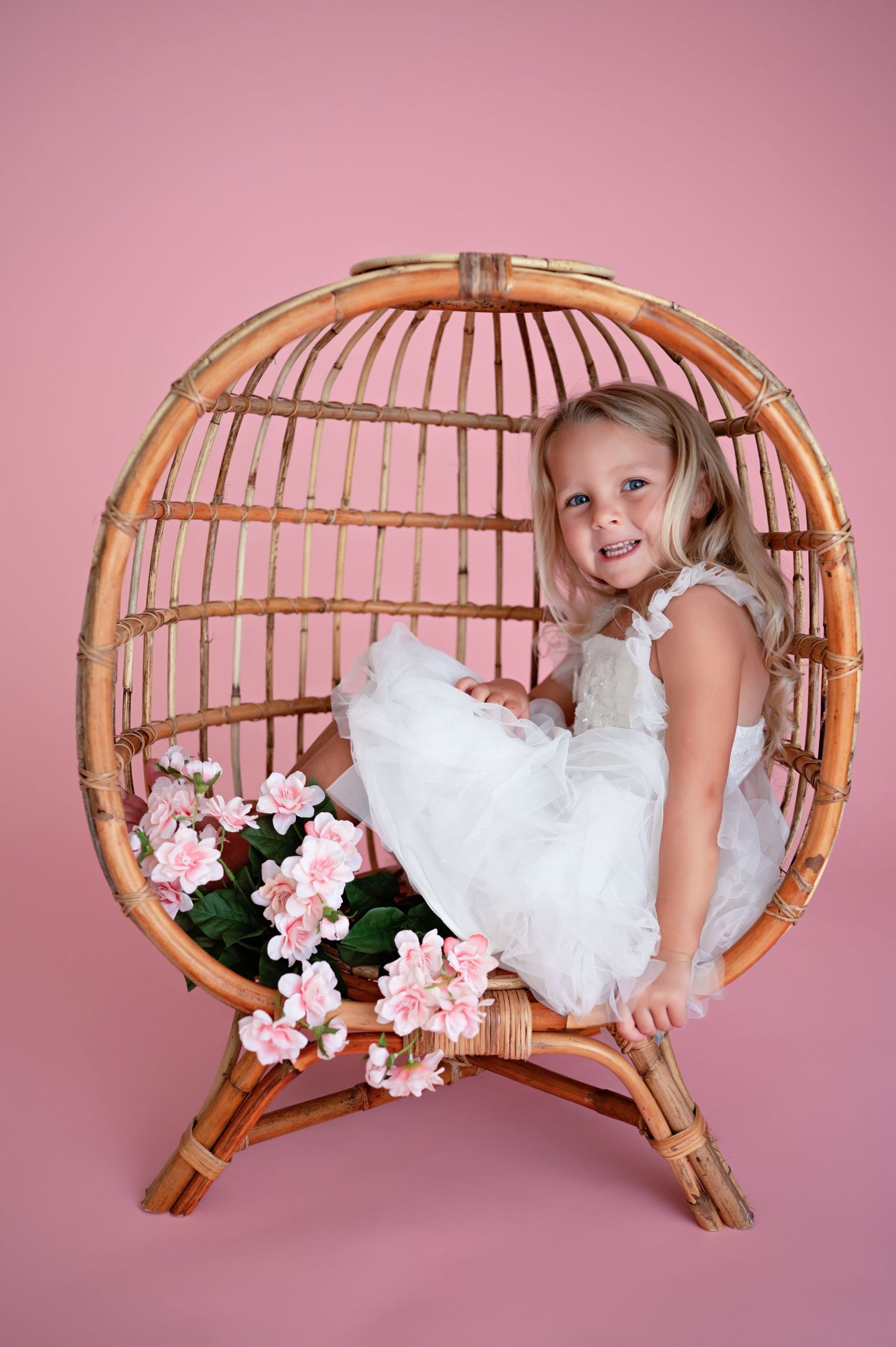 A decorative circular bamboo chair against a pink backdrop features a fluffy white dress and pink floral accents.