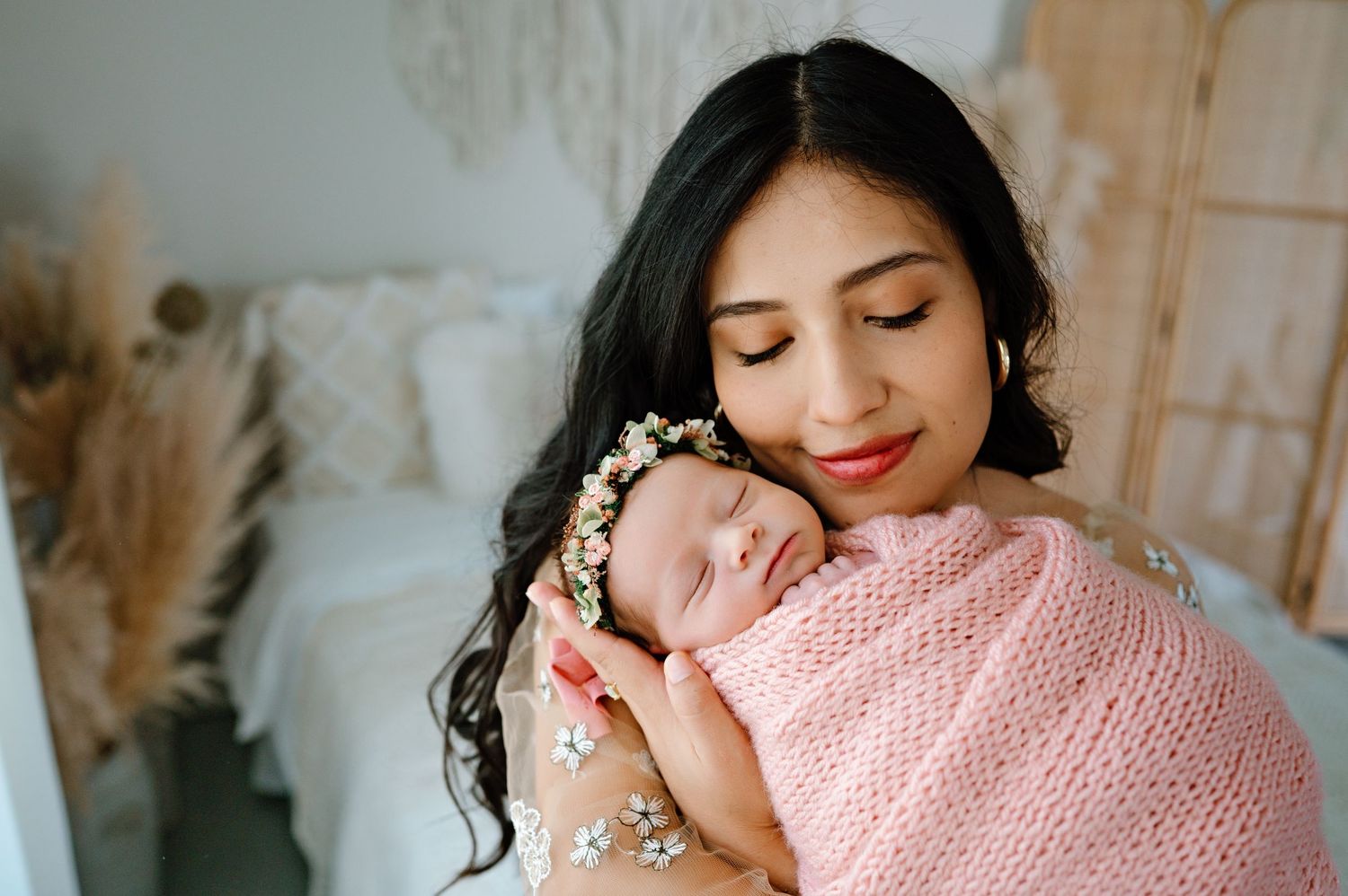 Woman and sleeping newborn in pink wrap surrounded by dried flowers in natural light studio setting.