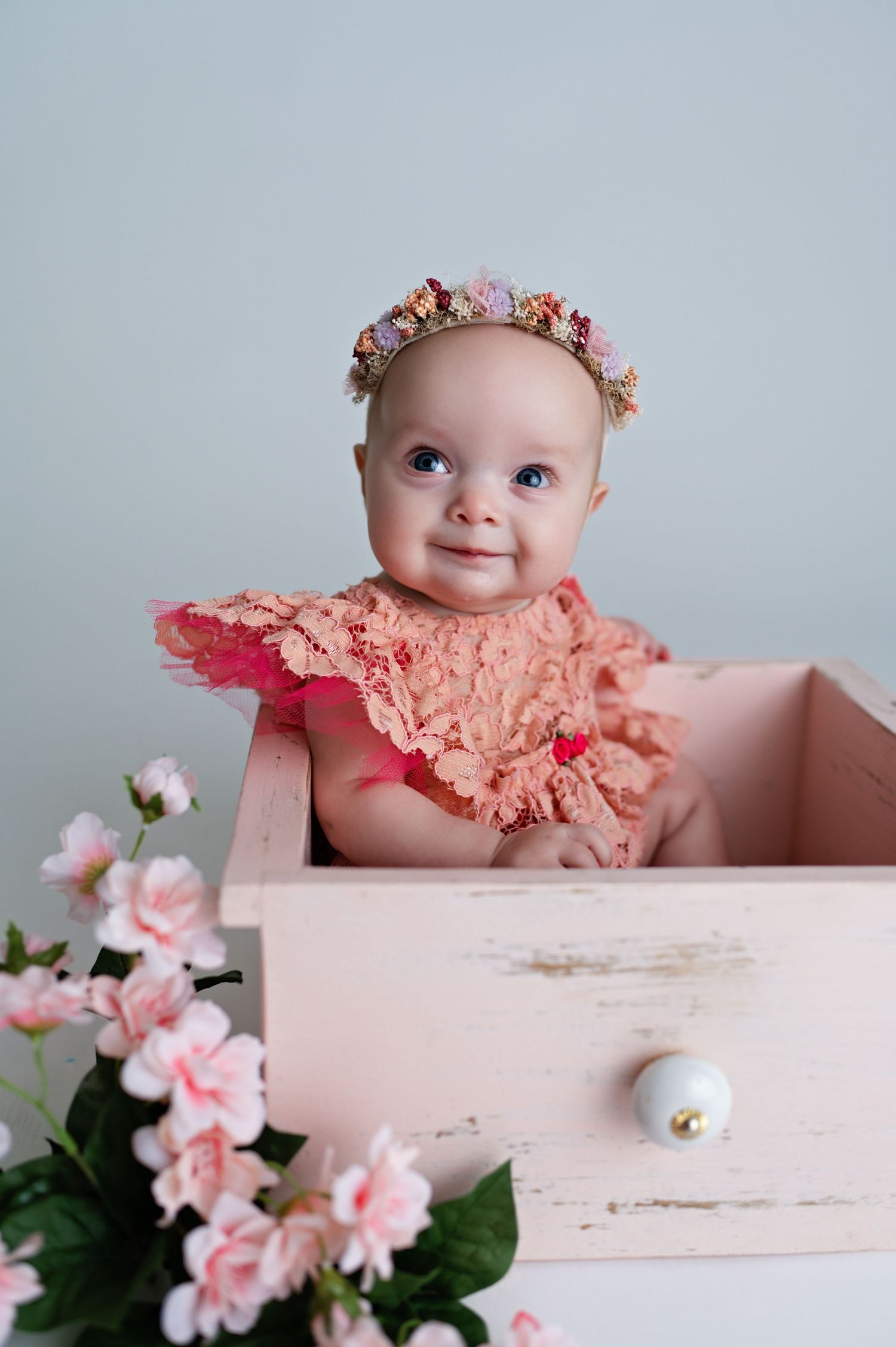 Baby in peach outfit sits in distressed white wooden box with pink flowers.