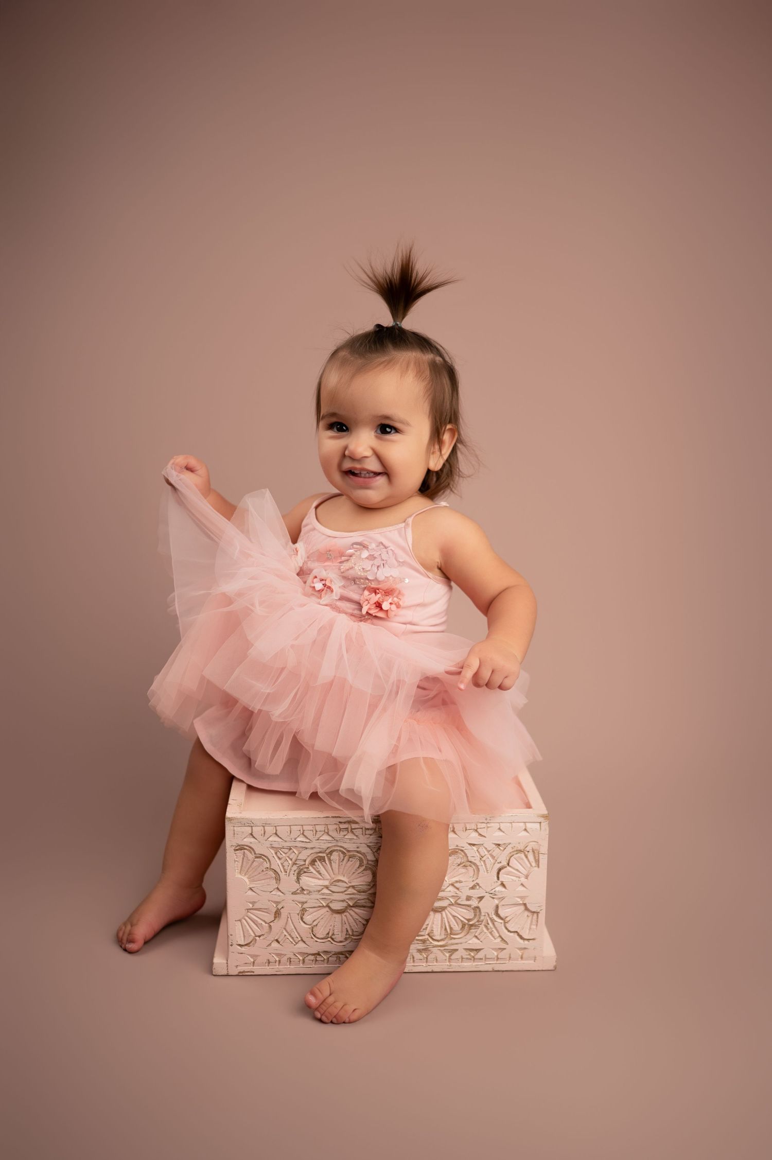 Baby wearing pink tulle dress poses playfully in ornate white wooden box during photo session.