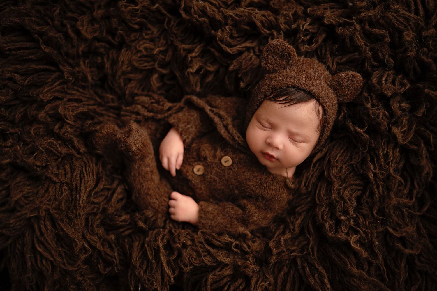 A sleeping newborn wearing a brown bear costume lies on a textured brown backdrop during a photography session.