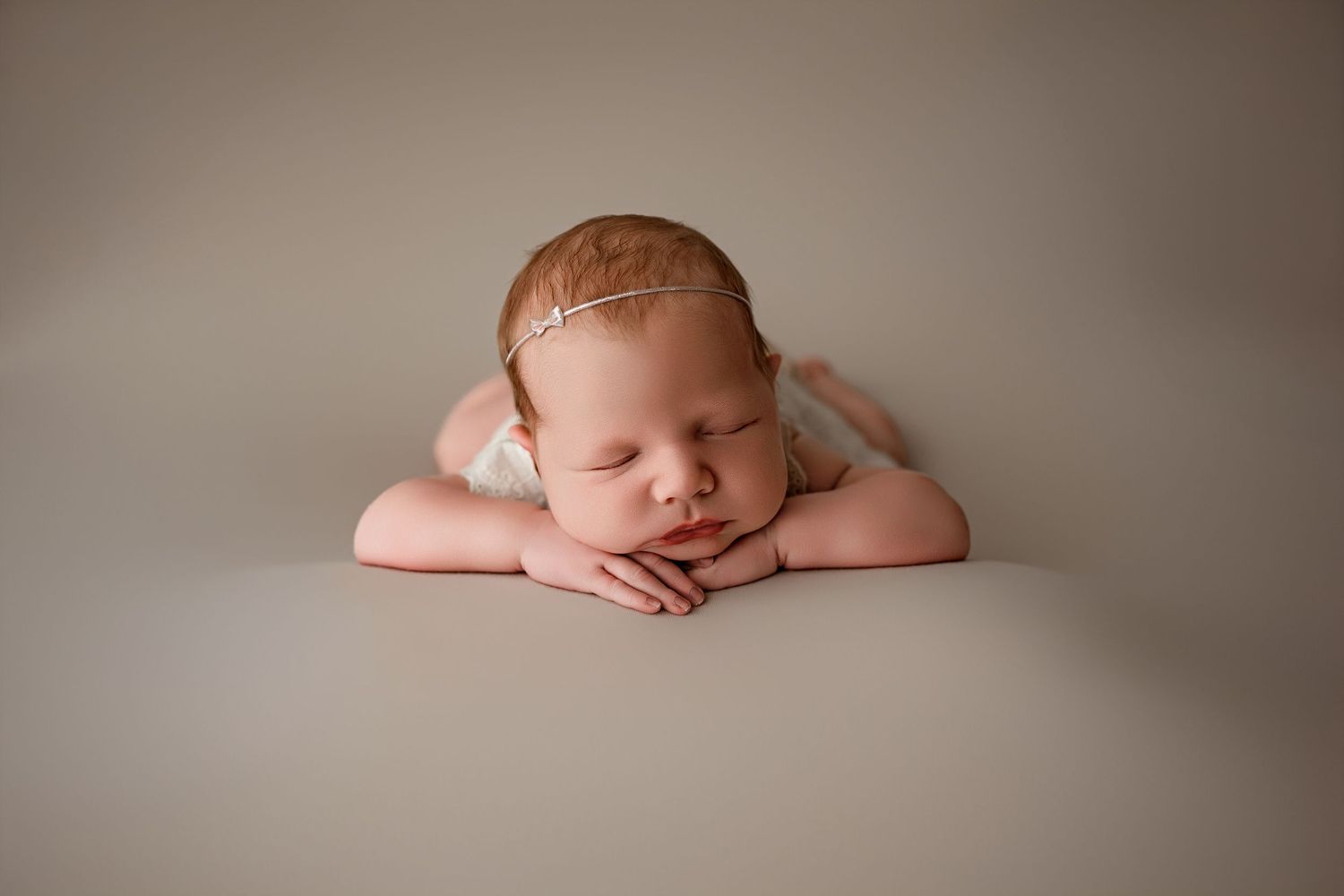 Sleeping newborn baby posed with hands under chin on neutral gray background wearing delicate gold headband.