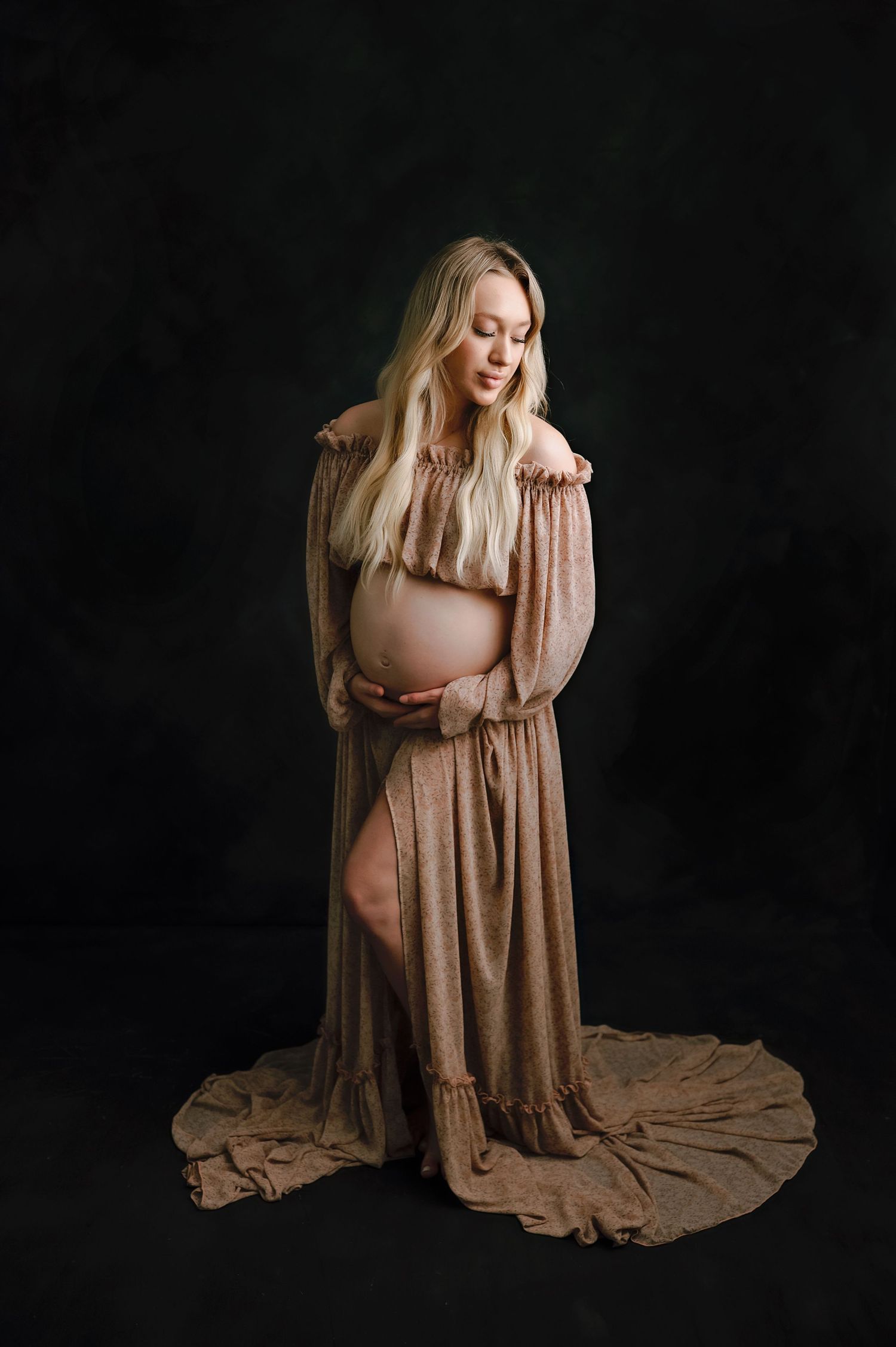 Maternity portrait featuring flowing tan dress against dark backdrop highlighting pregnant silhouette.