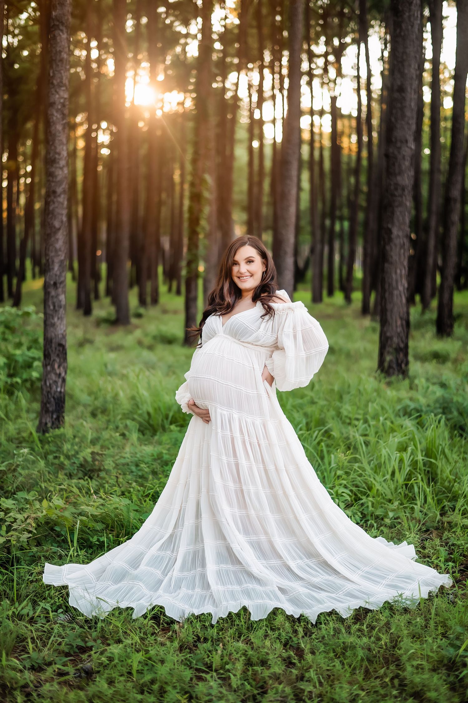 A maternity portrait in a flowing white dress among tall pine trees at sunset.