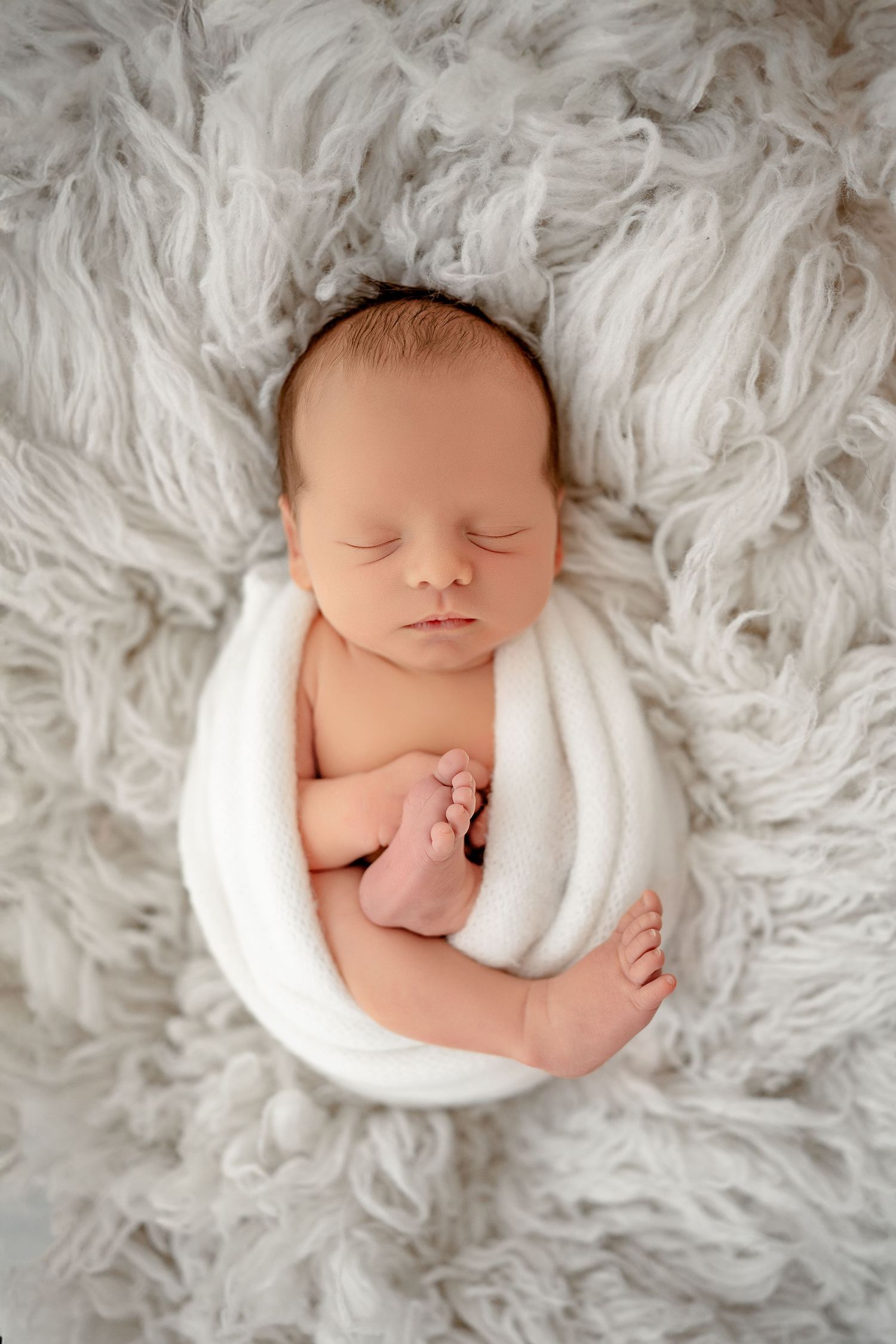 Sleeping newborn wrapped in white blanket nestled in soft fluffy white fur backdrop.