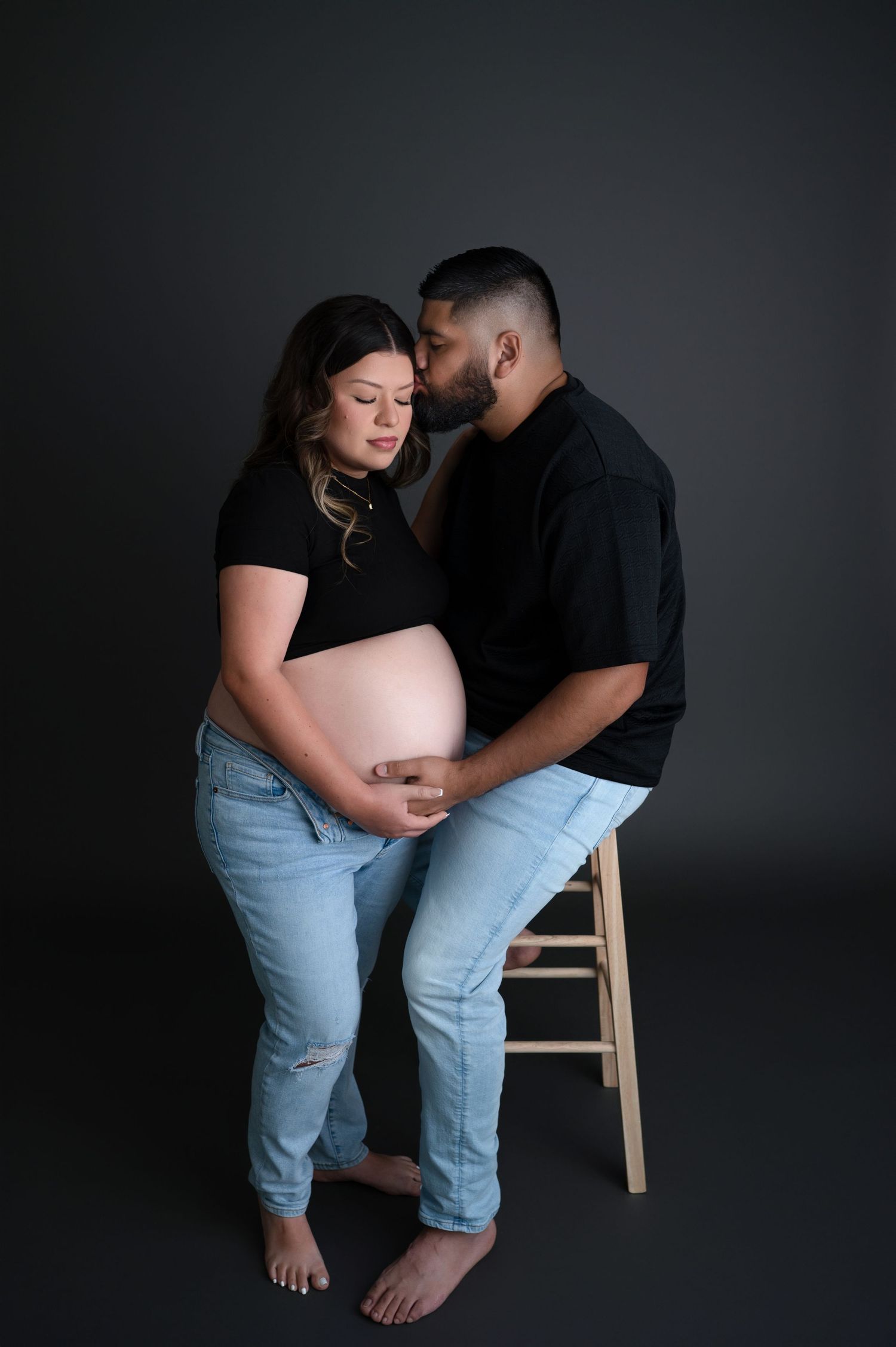 Intimate maternity photo of couple in black shirts and light jeans embracing against dark background.