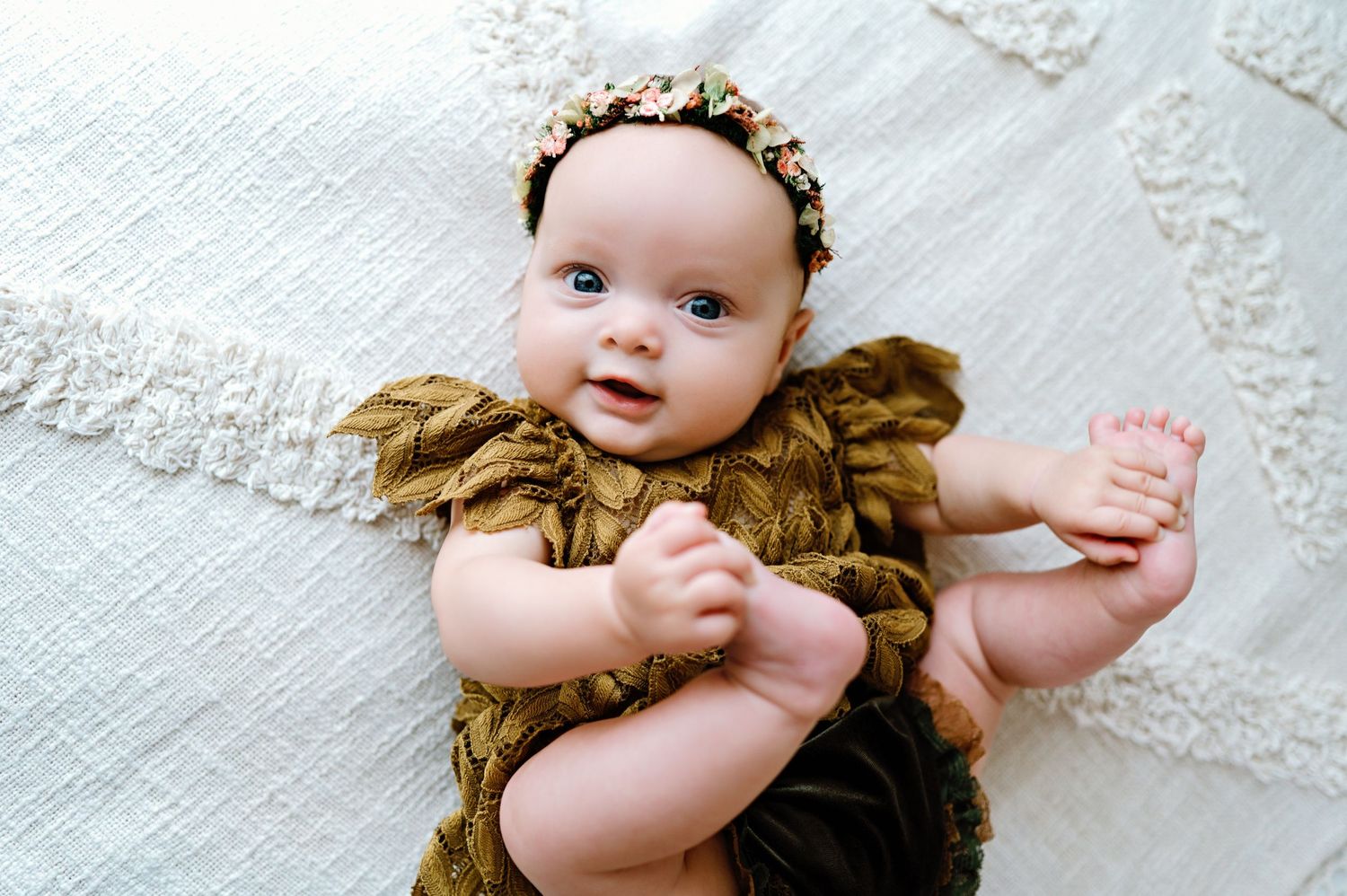 Small infant wearing golden ruffled dress with floral headband lies on white lace background.