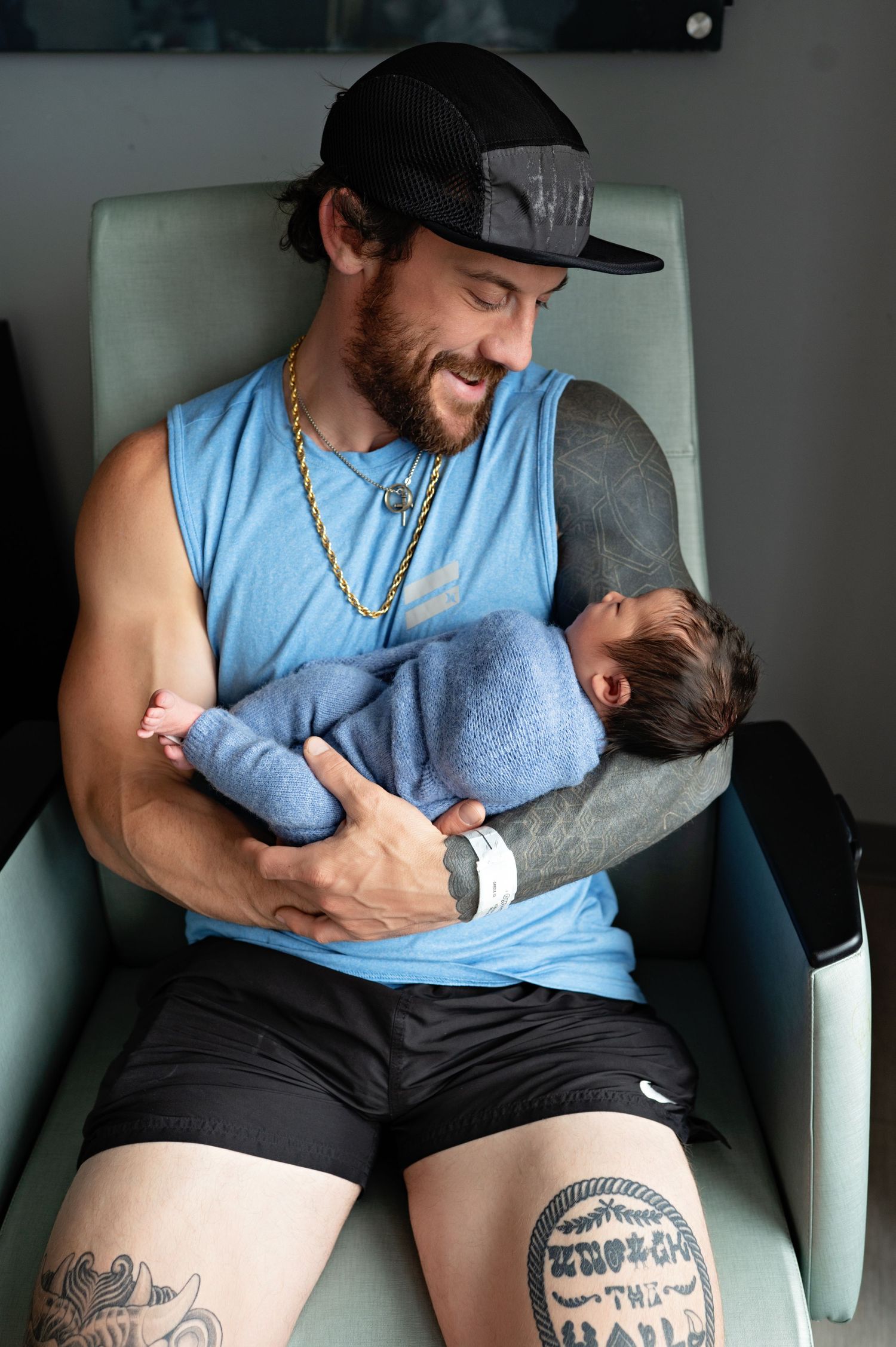A person in a light blue tank top and black shorts sits in a chair holding a sleeping baby in grey clothing.
