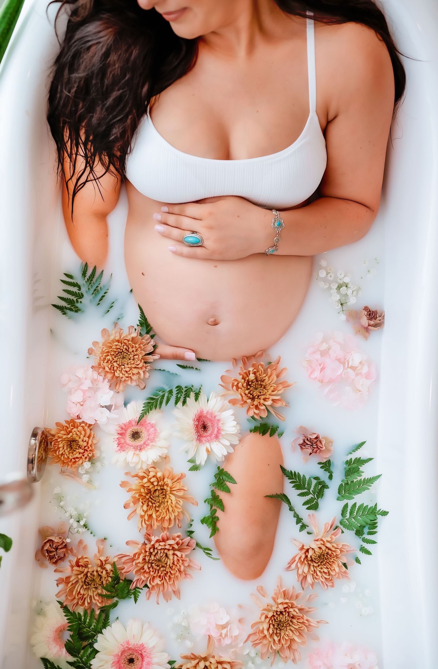 Top view of pregnant woman's belly surrounded by pink and orange flowers with green ferns in a milk bath setting.