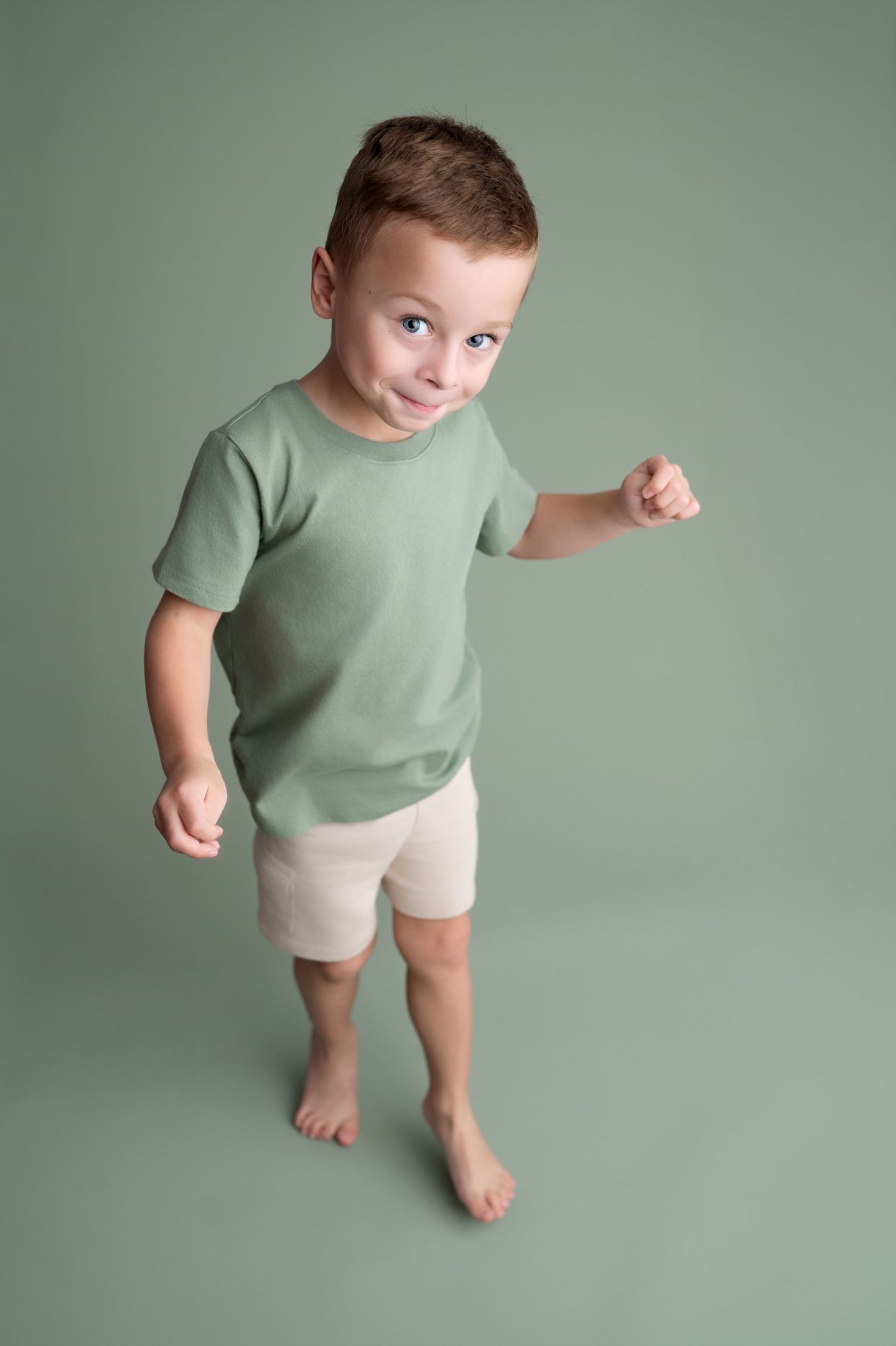 Young child in mint green t-shirt and beige shorts poses against sage green backdrop in studio setting.
