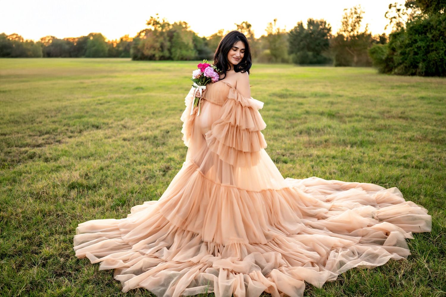 A flowing peach-colored tiered gown with a dramatic train spreads across a grassy field at sunset.