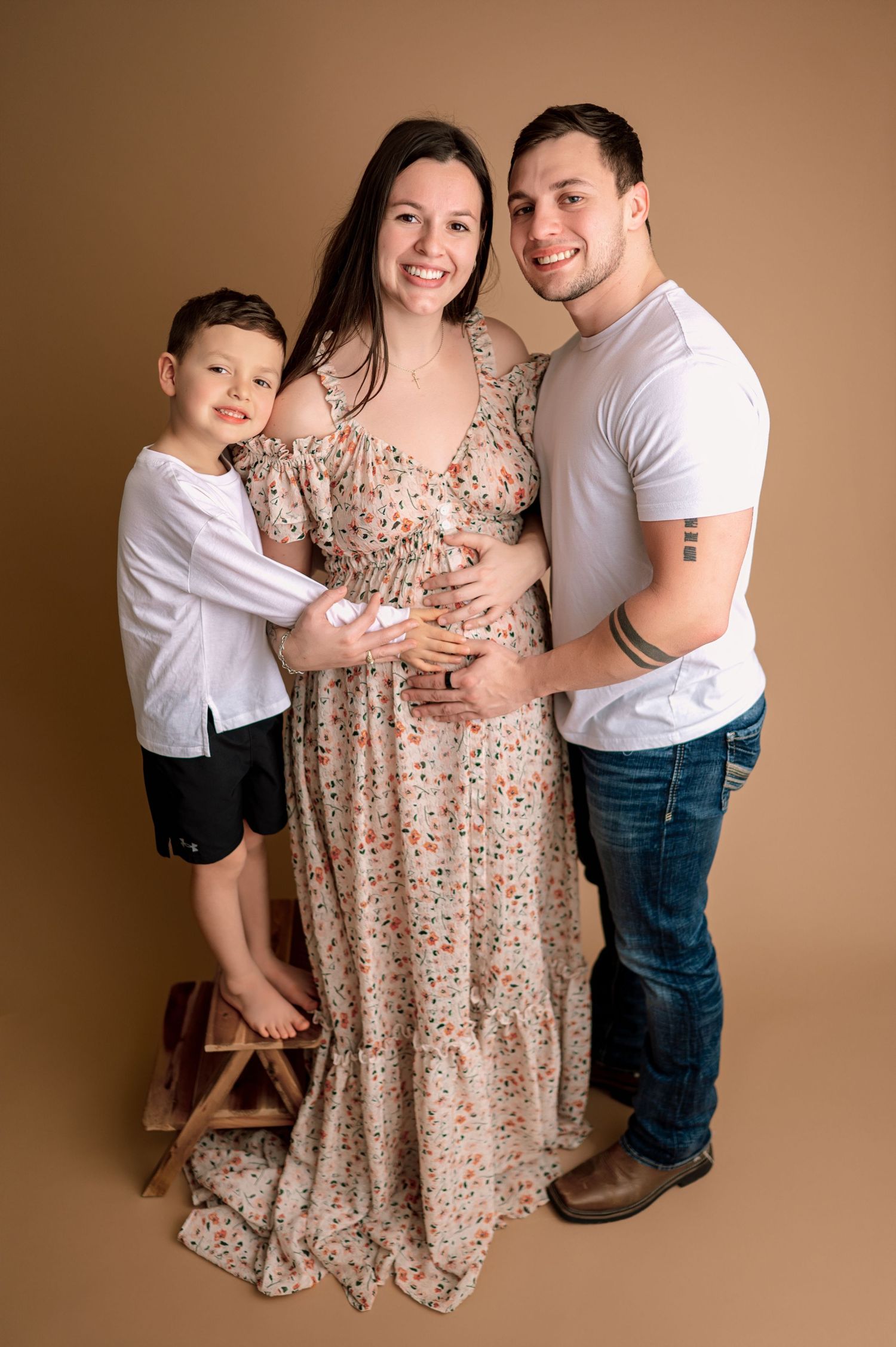 Maternity photo series showing a pregnant woman in floral dress with family members posing against brown backdrop.