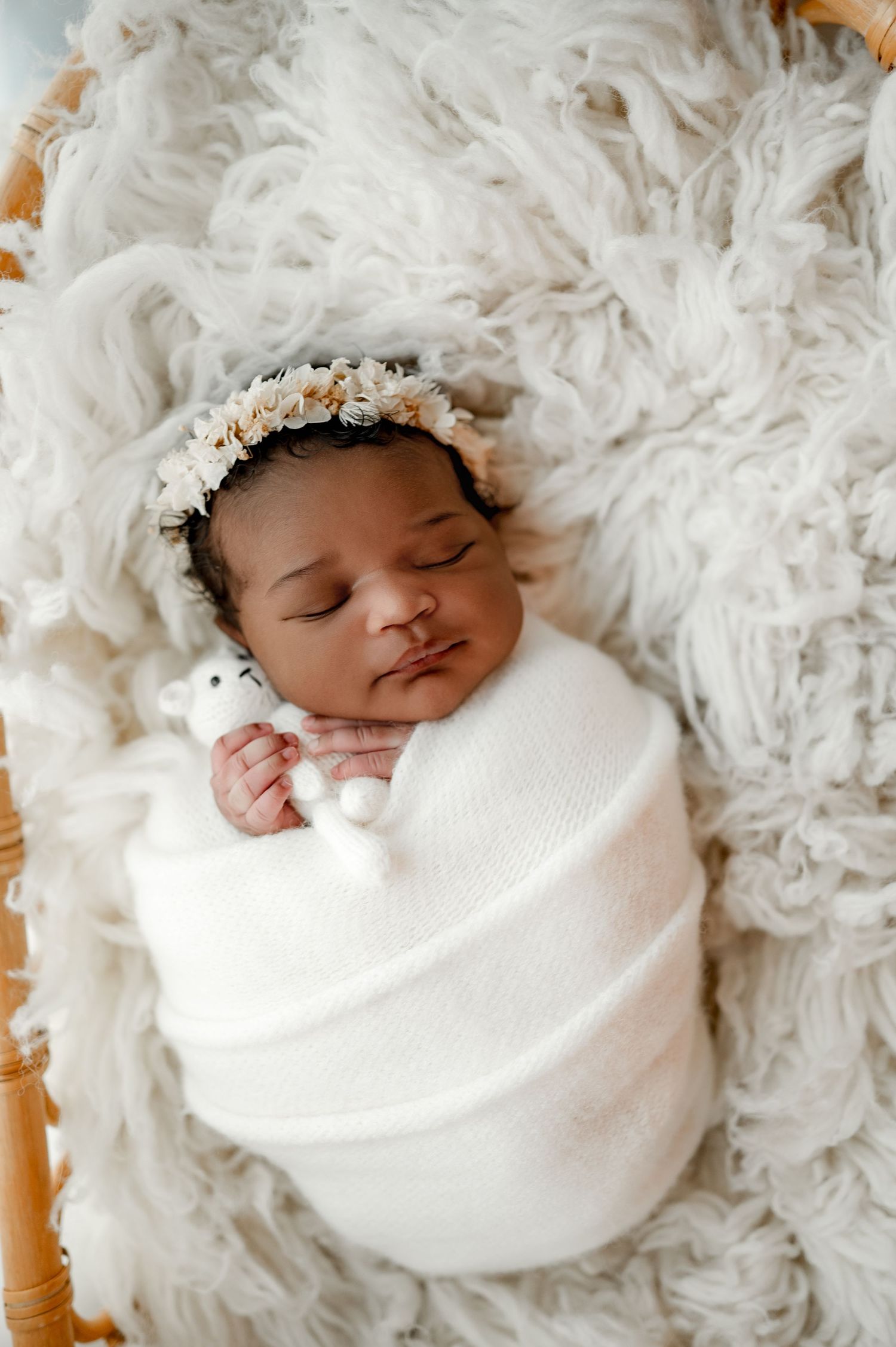 Sleeping newborn swaddled in white wrap with floral crown lying on soft fluffy white textured blanket.