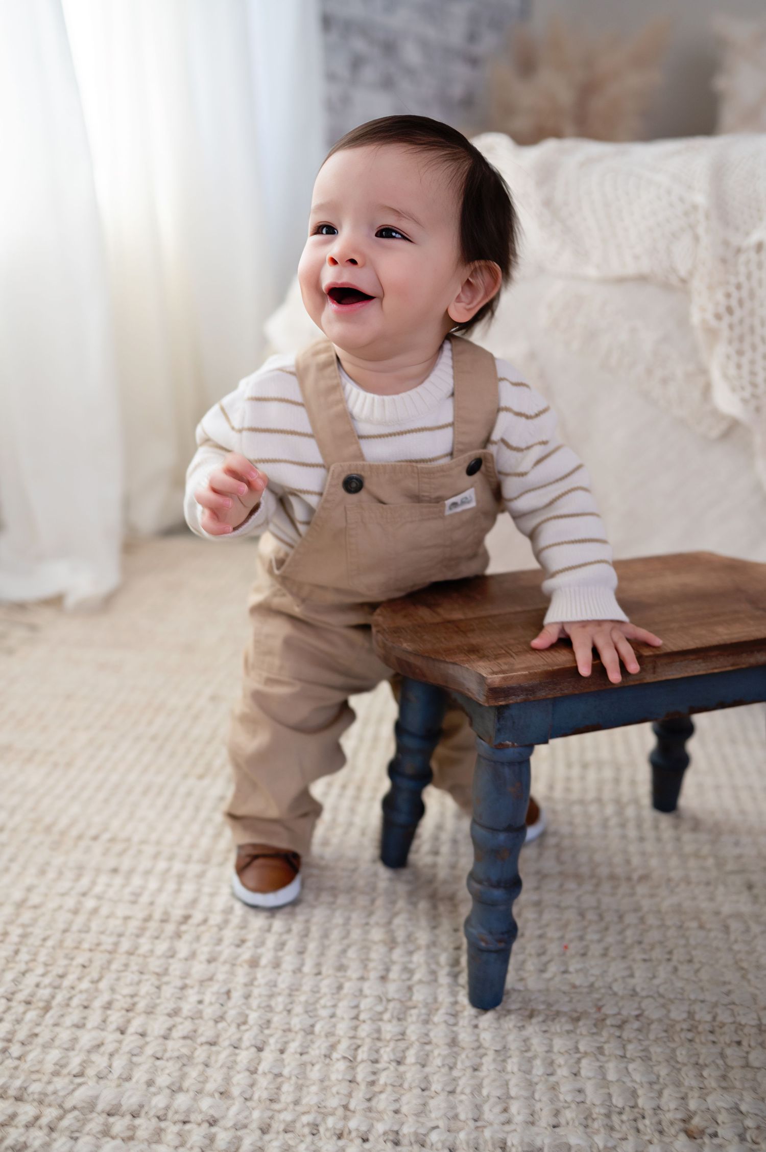A laughing baby in tan overalls and striped shirt stands holding onto a blue wooden bench on a beige carpet.