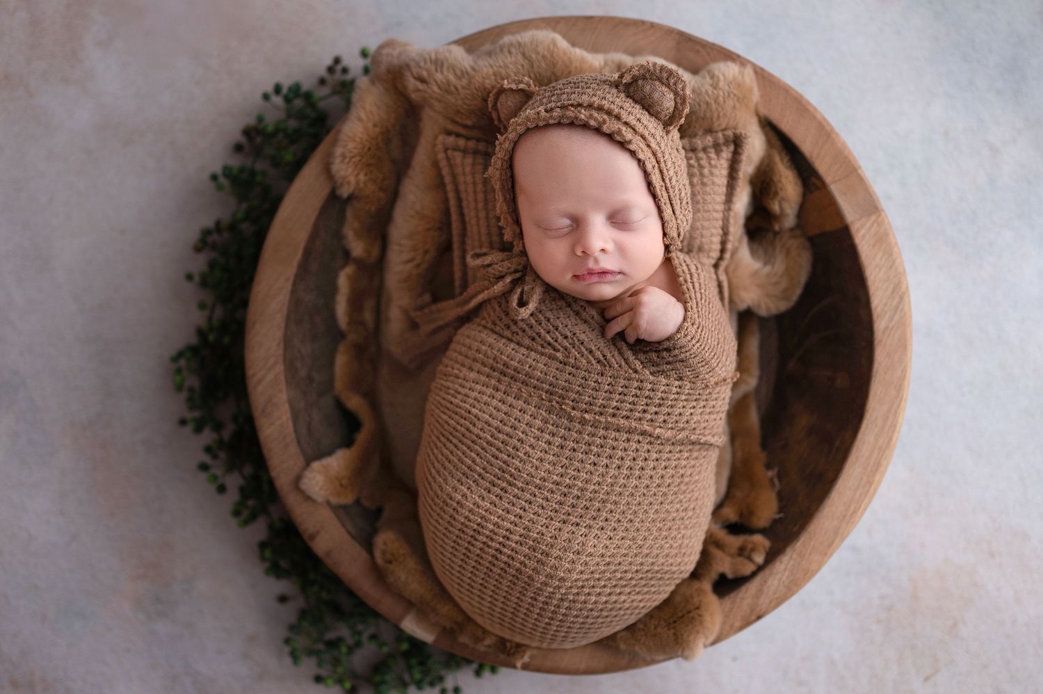 Newborn baby wrapped in cozy brown knit blanket sleeping peacefully in rustic wooden bowl with greenery accent.