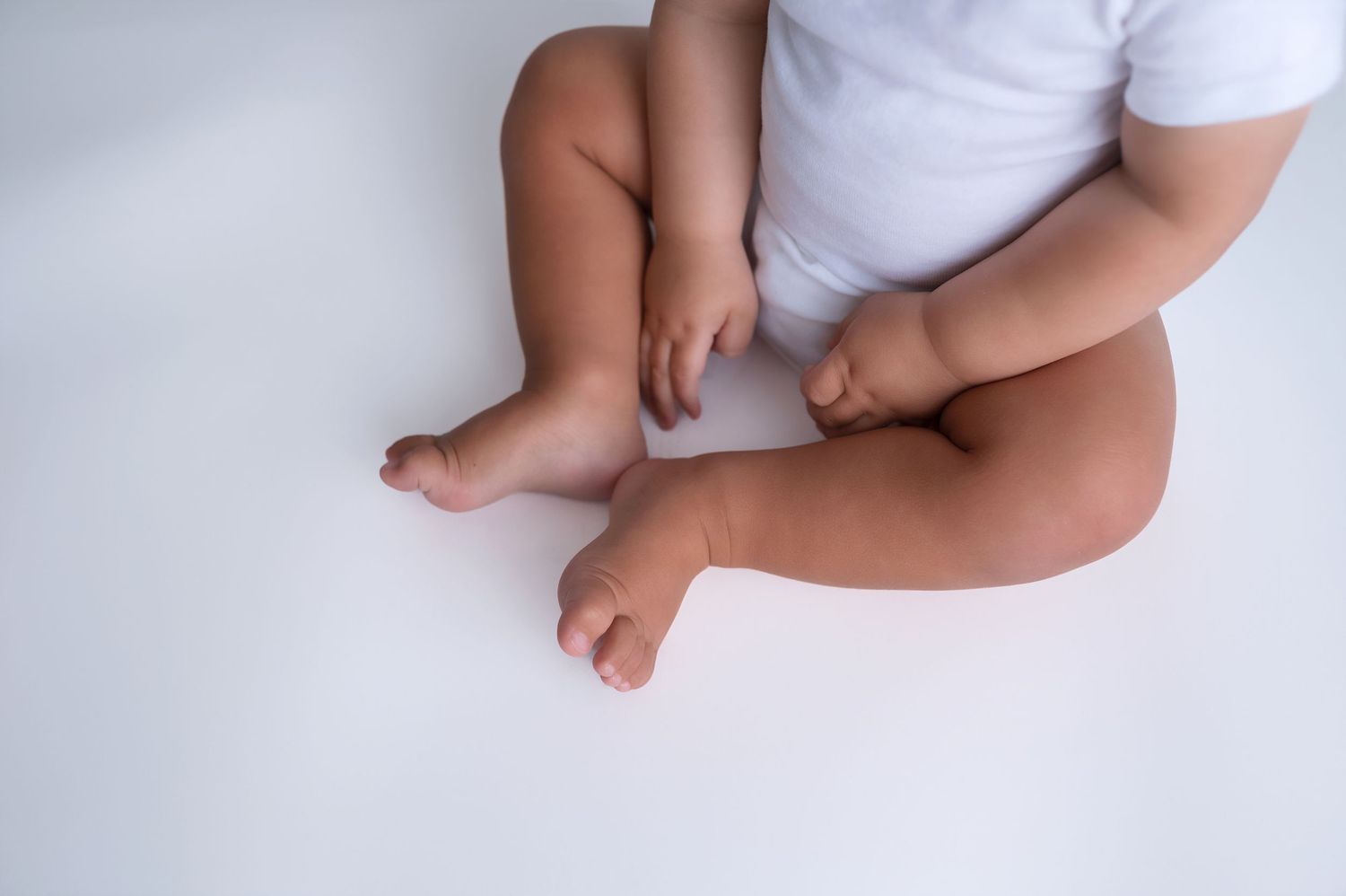 Close-up of baby's feet and legs in white onesie against light background.