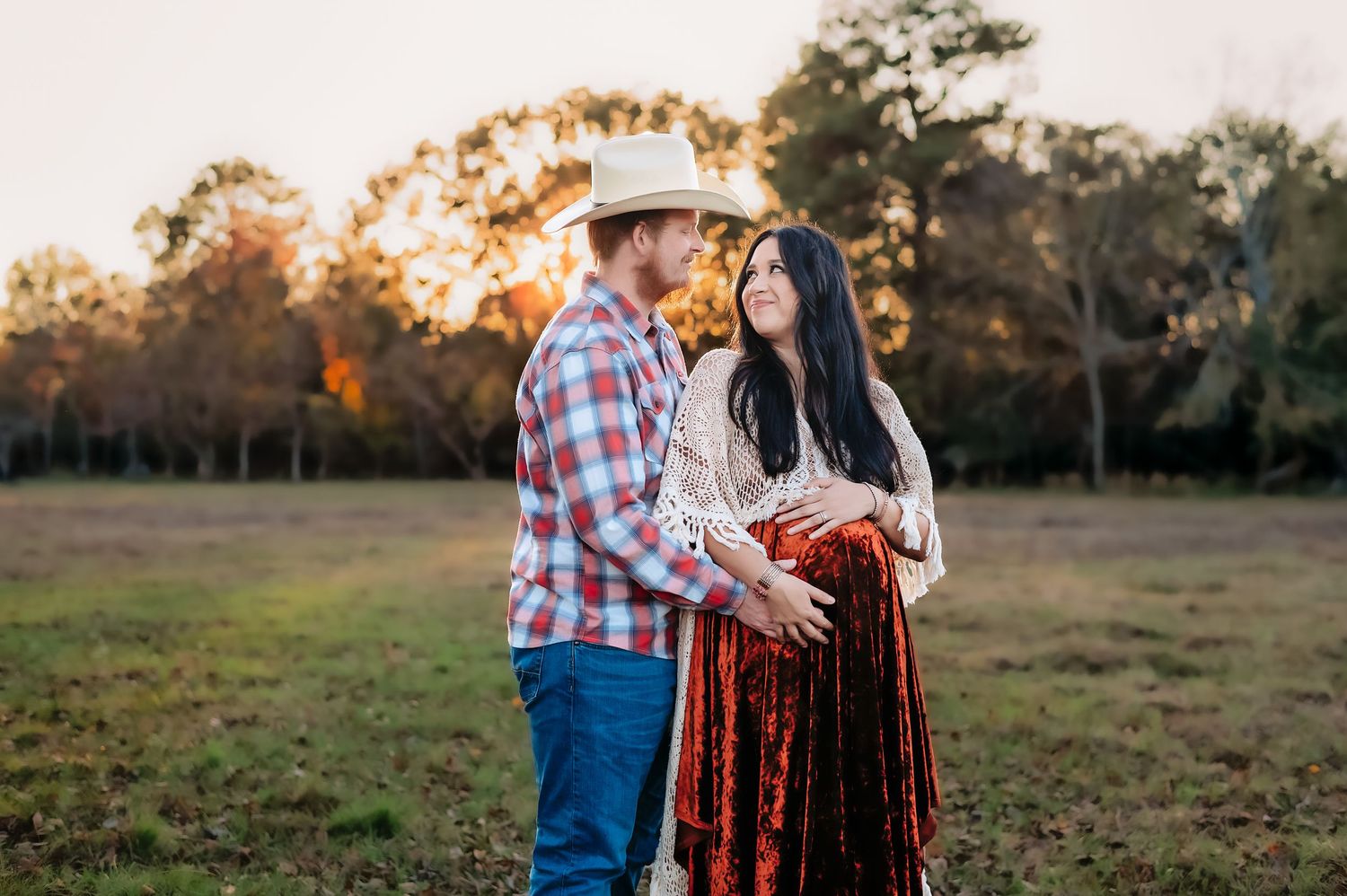 A couple poses together in a country field at sunset with a rustic barn in the background.