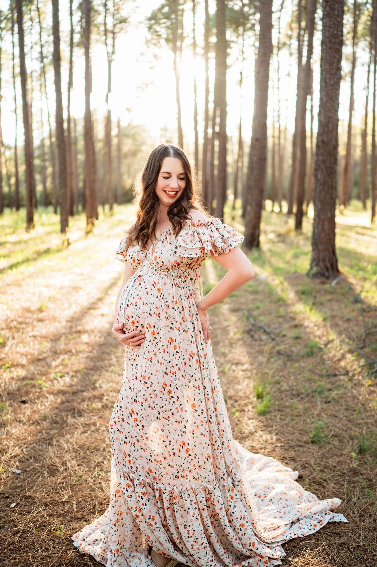 Pregnant woman in floral maxi dress posing in sunlit pine forest for maternity photo session.