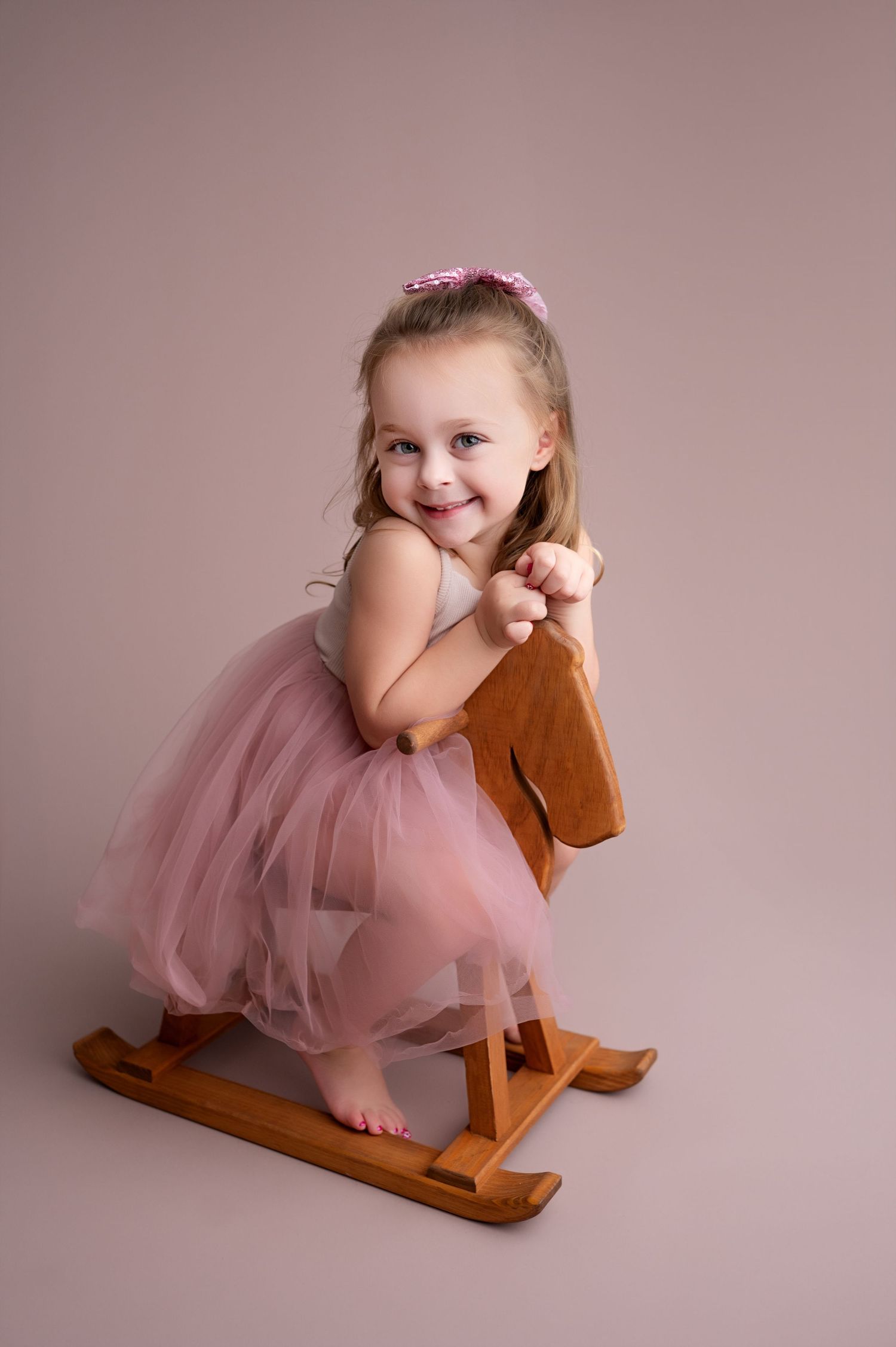 Child in pink tulle skirt sits on wooden rocking horse against mauve backdrop.