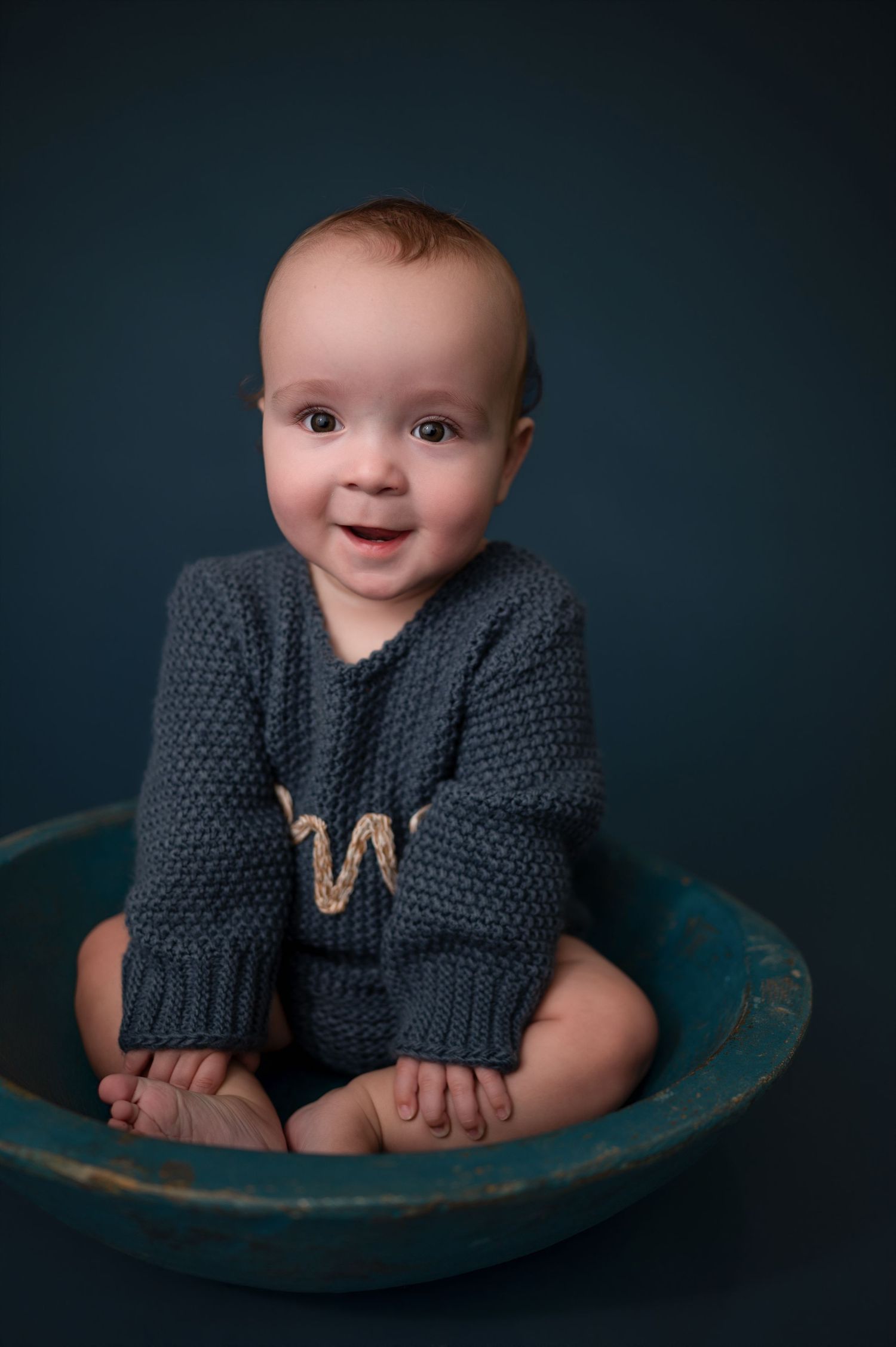 A small baby wearing a gray knit cardigan sits in a vintage teal bowl against a dark background.