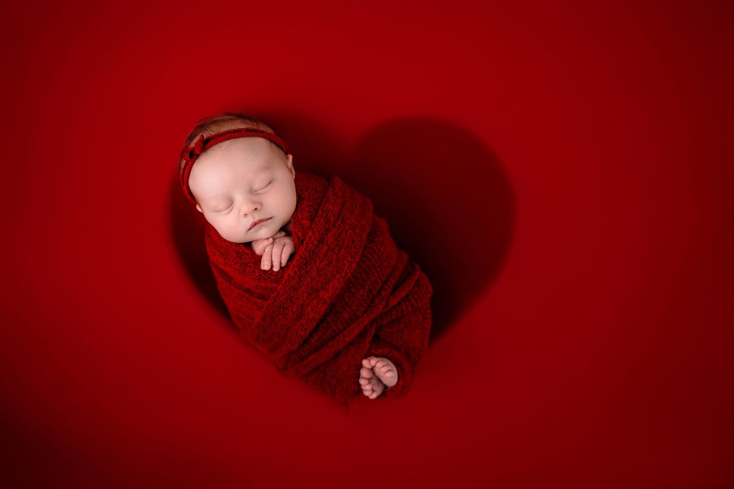 Sleeping newborn baby wrapped in red swaddle blanket on red background creating heart-shaped shadow.