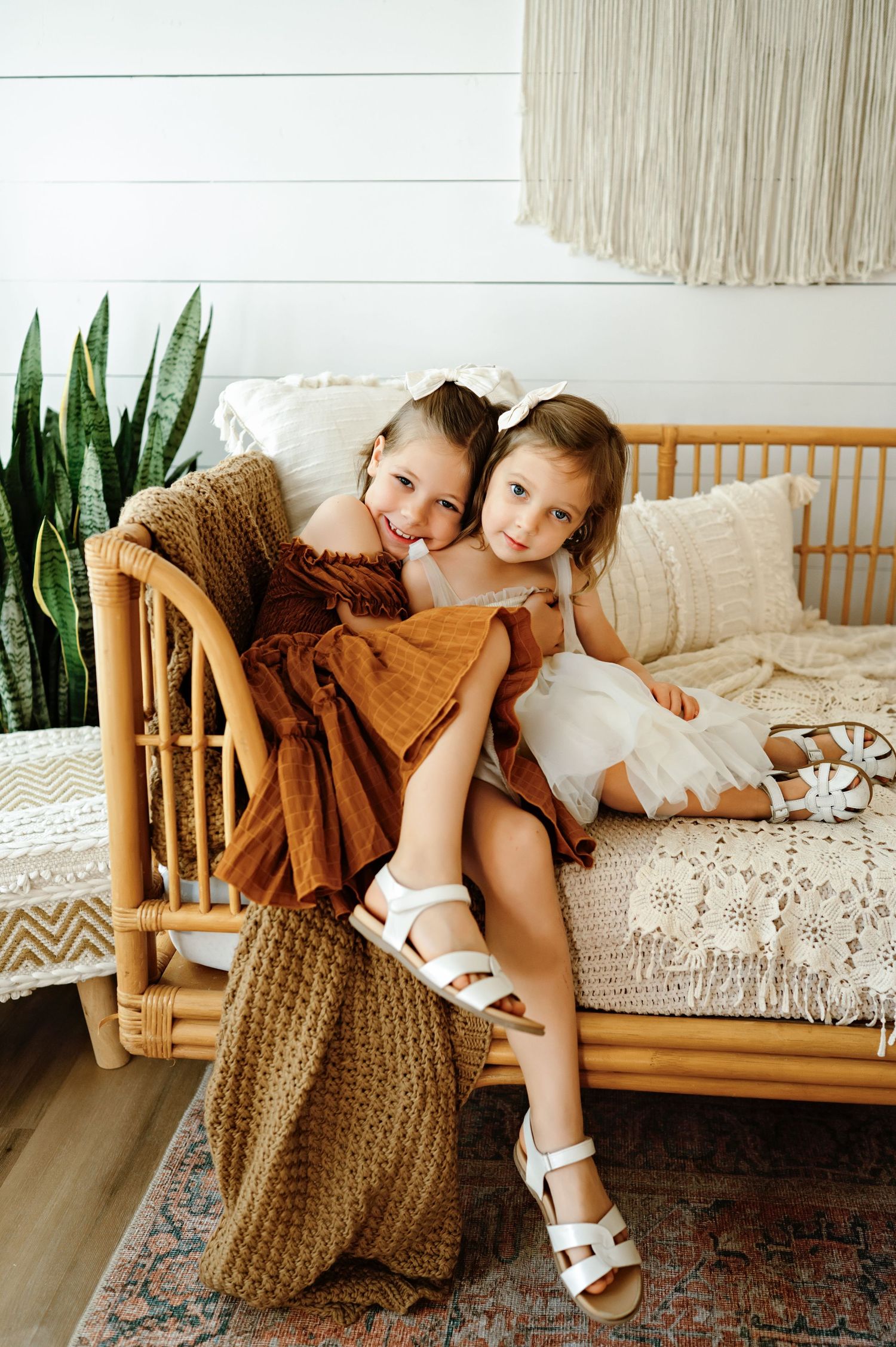 Two children relax together on a wooden daybed with brown cushions and decorative plants in a bright interior setting.