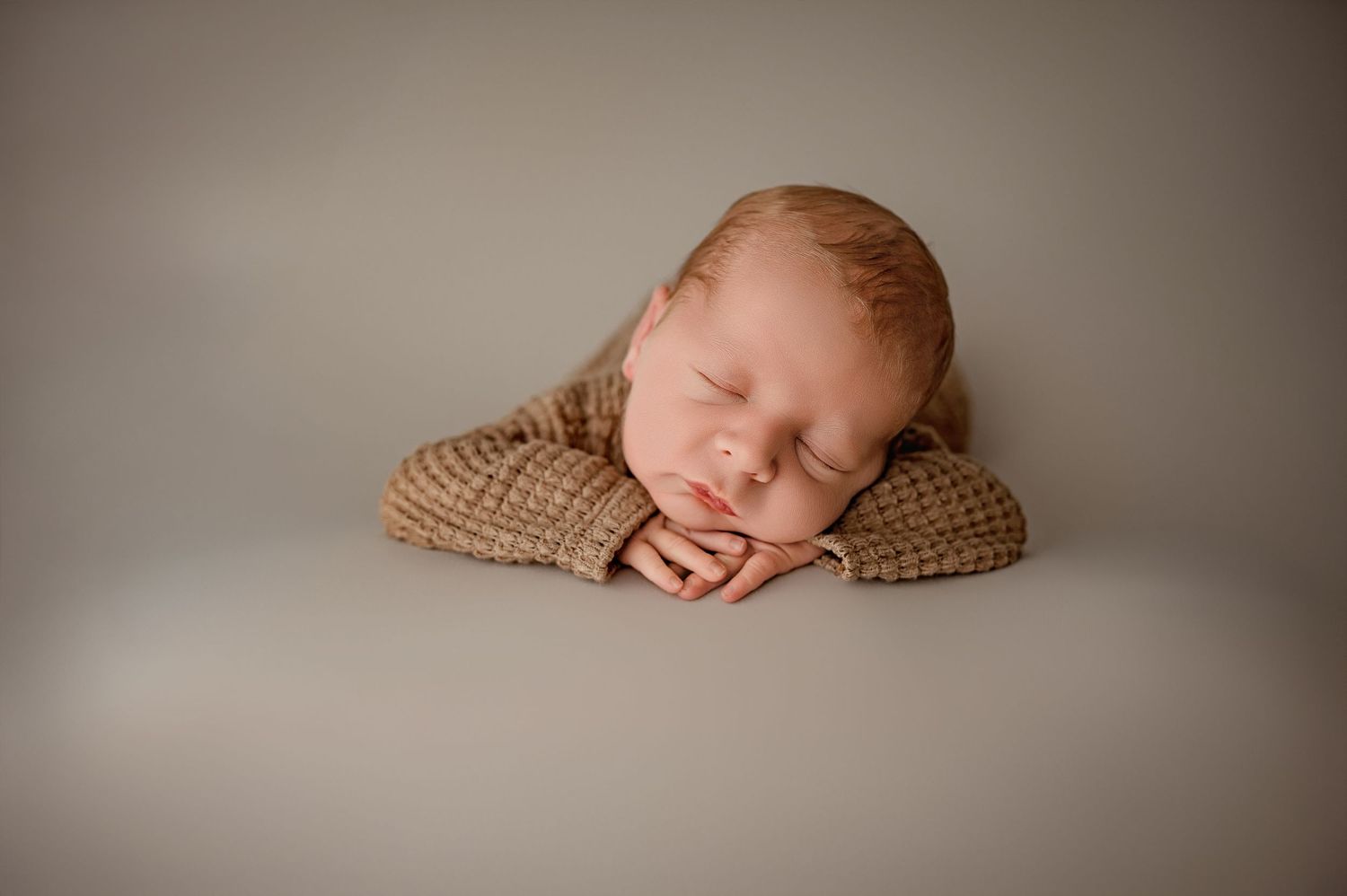 A sleeping newborn rests peacefully on a brown knitted blanket against a neutral gray background.