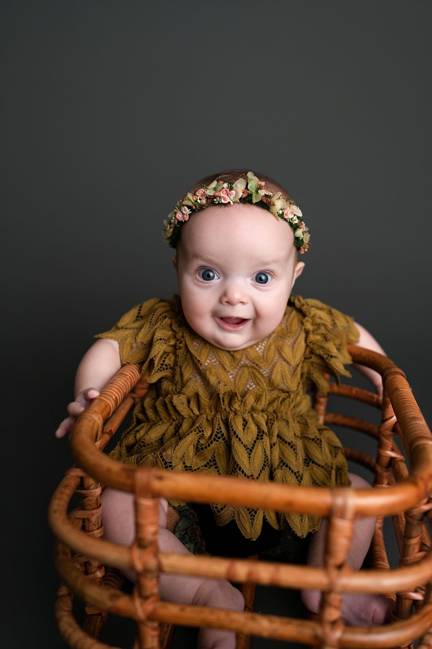 Baby in antique wood chair wearing mustard color ruffled outfit and floral crown on dark backdrop.