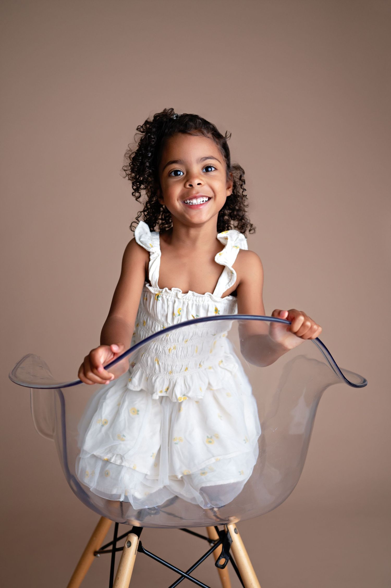 A child in a white dress sits playfully in a clear designer chair against a neutral background.