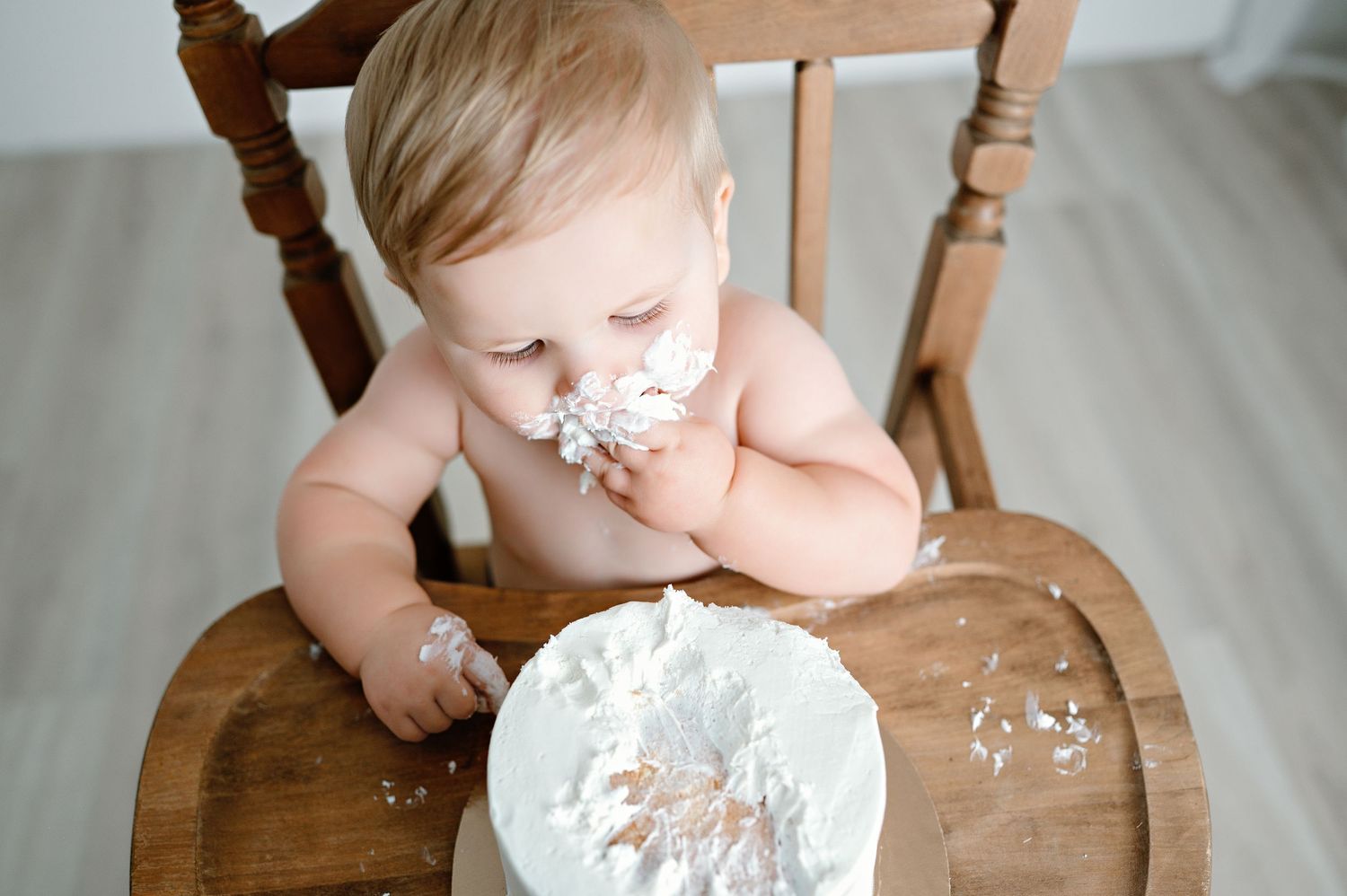 Baby sits in wooden high chair with cake frosting messily smeared on face and plate on tray.