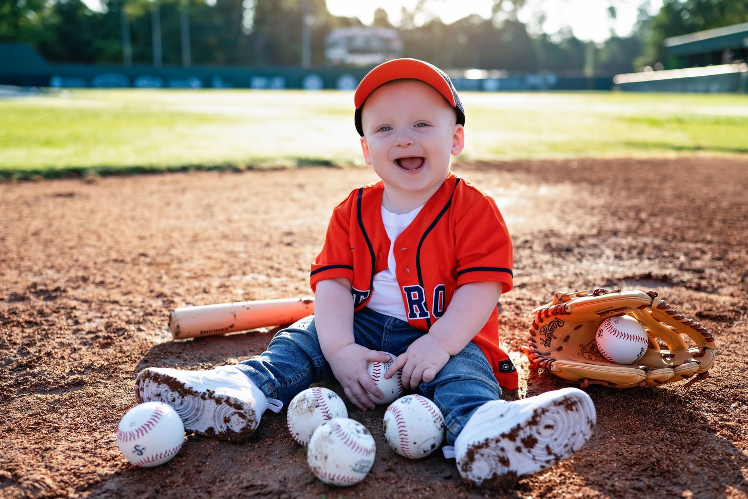 Small baseball player sitting on base path in red and white uniform smiling with ball field in background.
