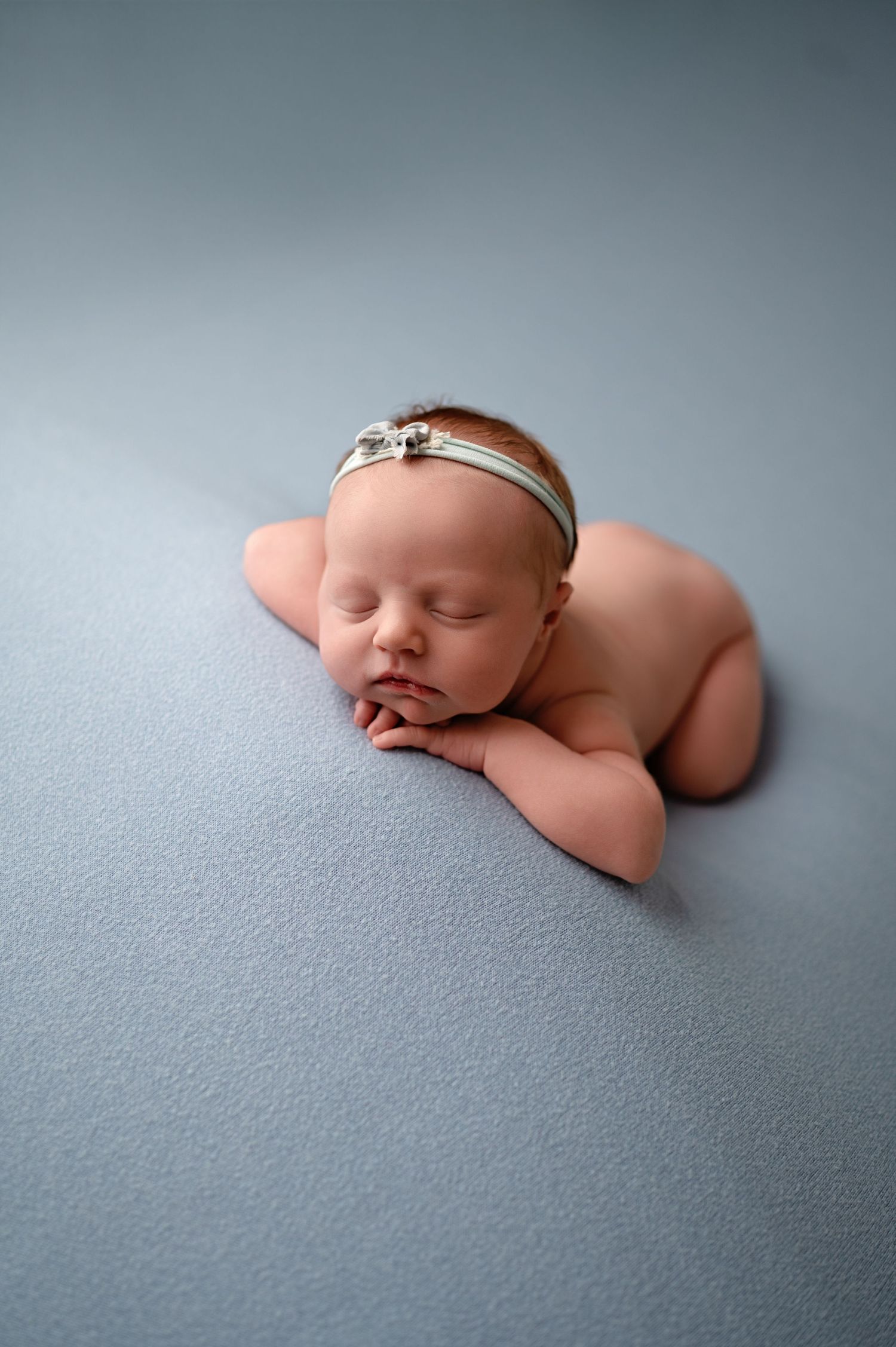 A sleeping newborn baby wearing a delicate headband lies peacefully on soft gray fabric in artistic portrait style.