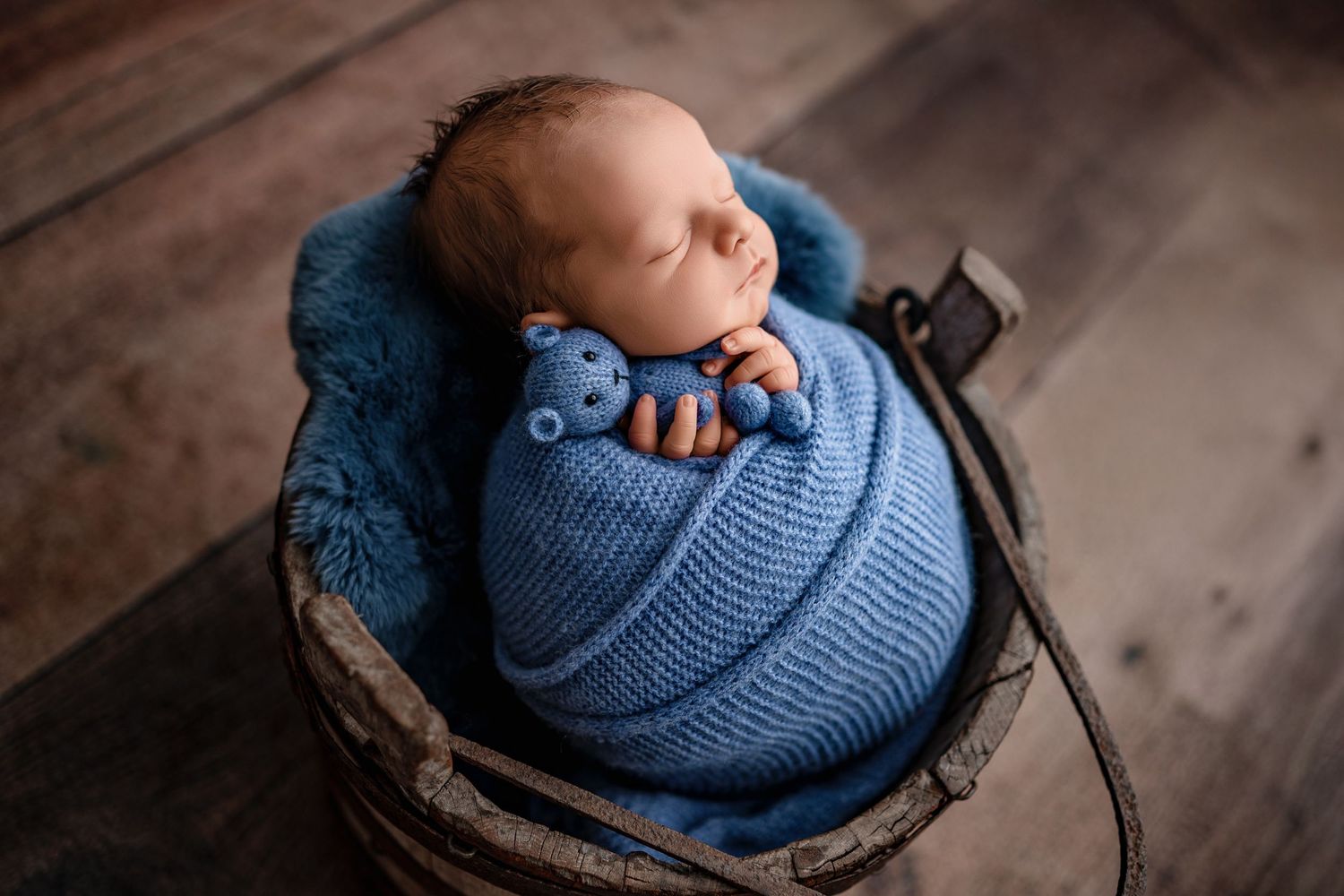 A baby wearing a blue knit sweater rests peacefully in a rustic wooden basket on a wooden floor.