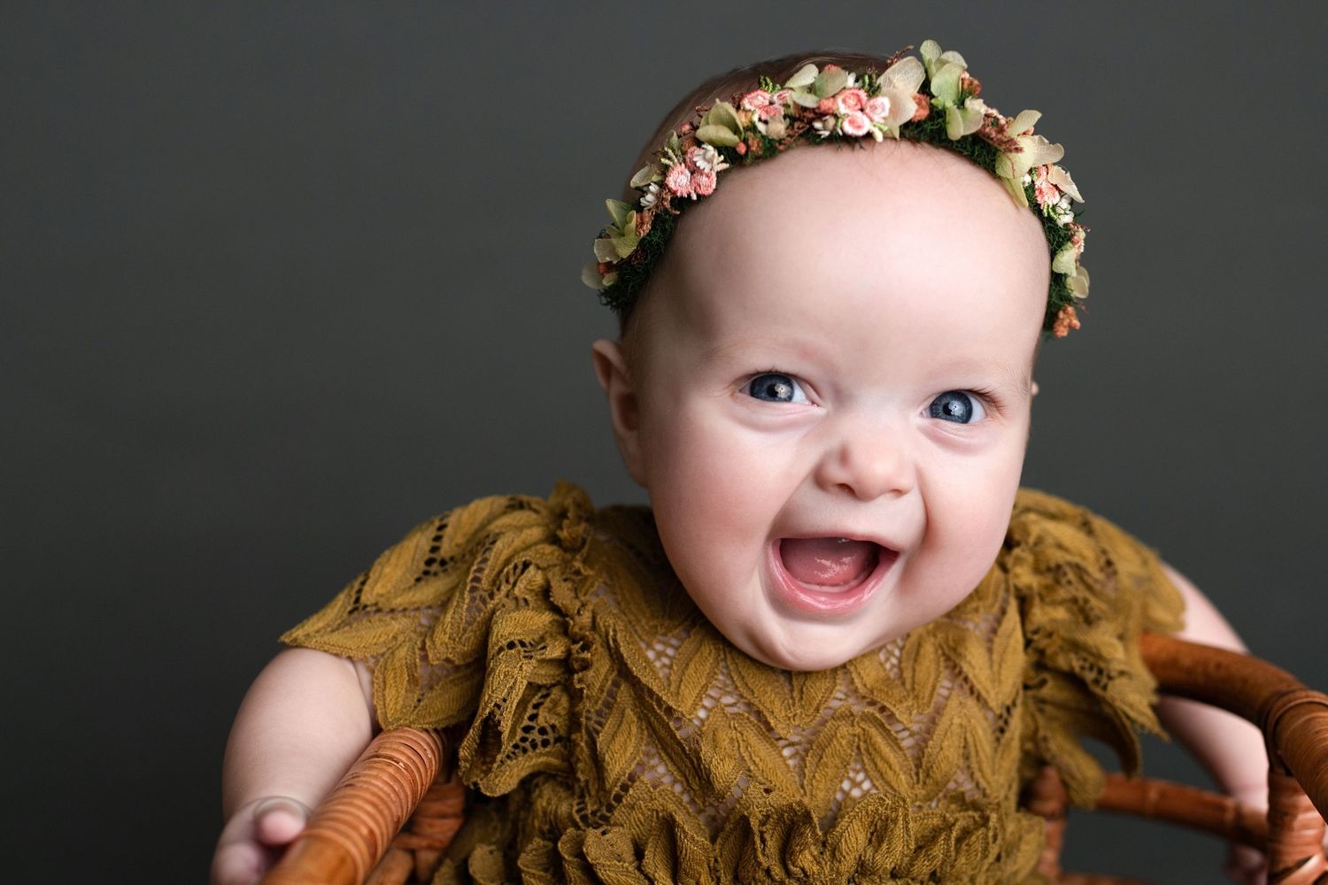 Happy baby wearing brown sweater and floral crown against dark background.