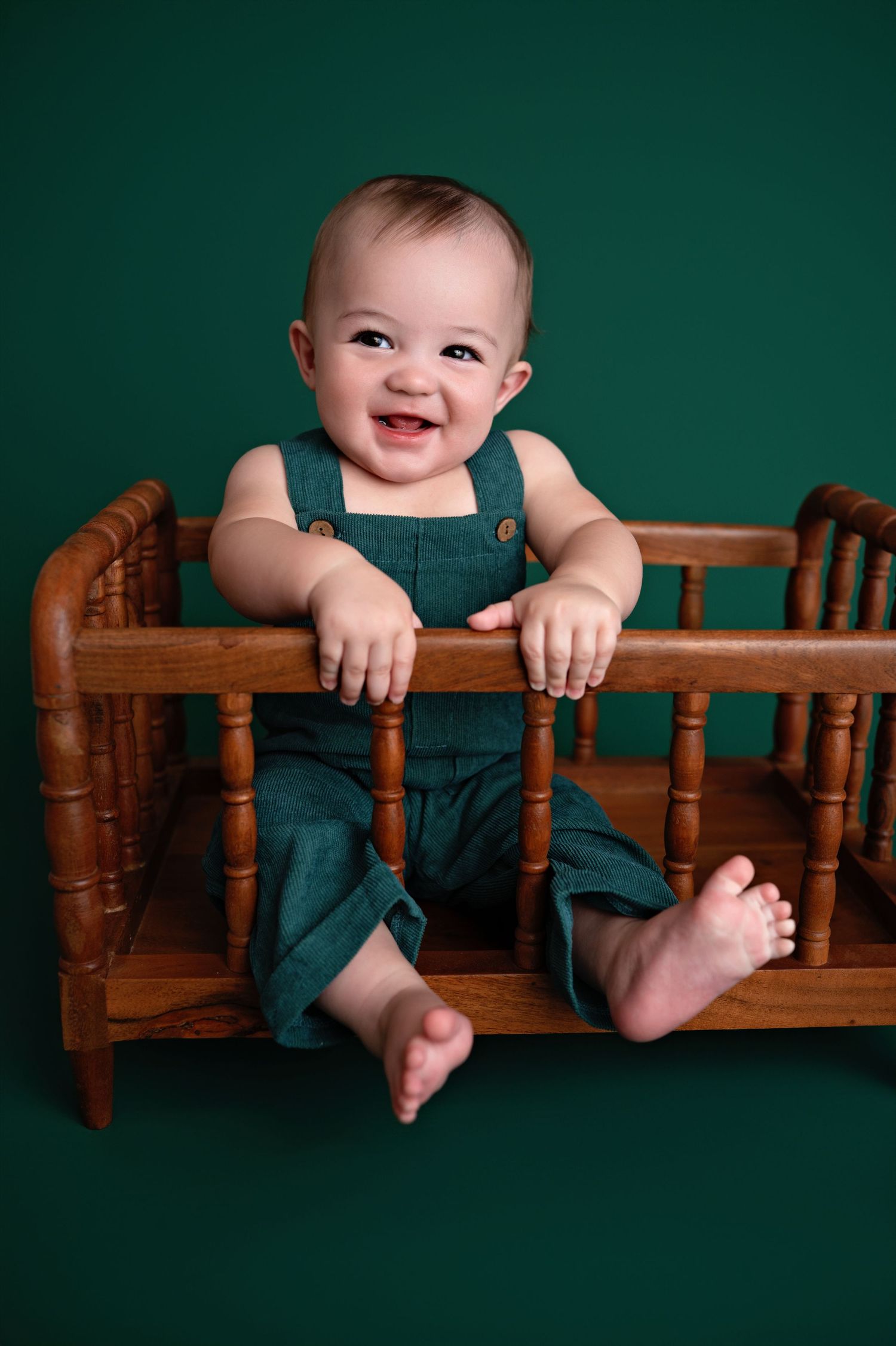 Baby in blue overalls sits behind wooden railing against dark green background in sequential poses.