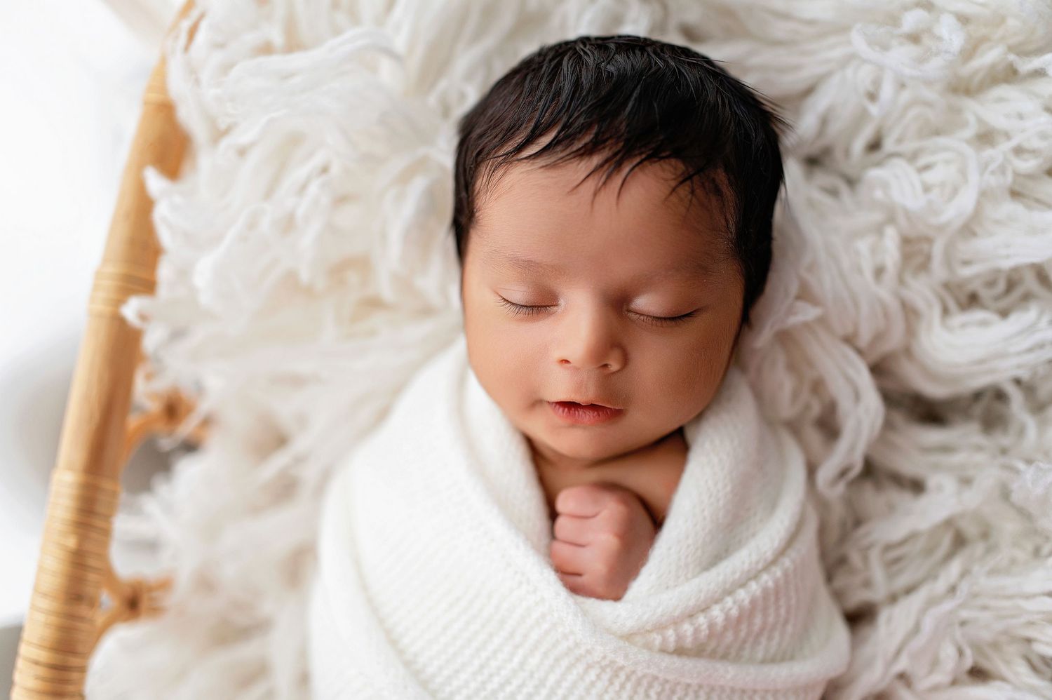 Sleeping newborn baby wrapped in soft white knit blanket lying on textured white backdrop.