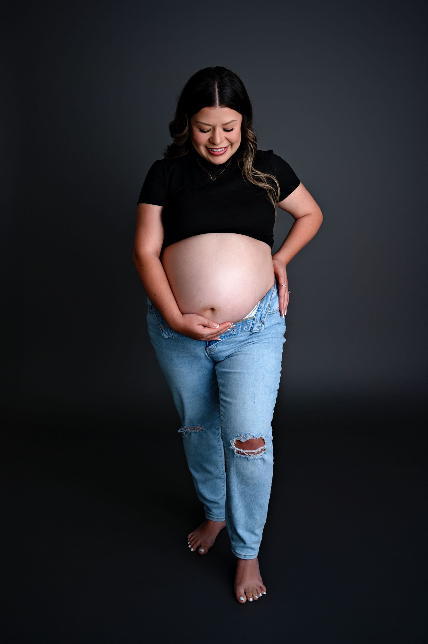 Maternity photo of an expectant mother in black shirt and ripped jeans against dark backdrop, looking down at her belly.