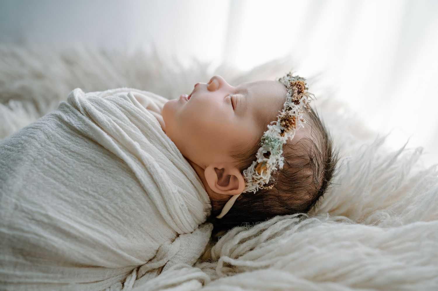A sleeping baby wearing a floral crown lies wrapped in white fabric on soft furry texture.