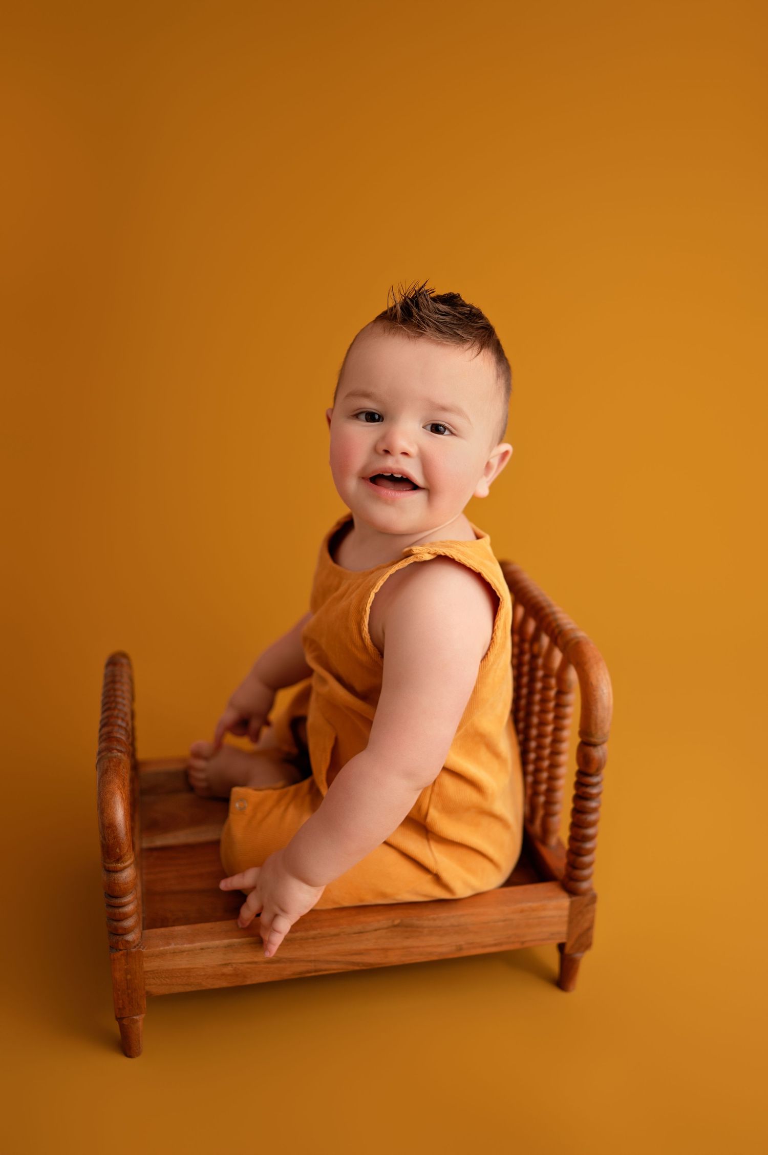 Baby in yellow onesie poses playfully on wooden doll bed against orange backdrop in professional photo series.