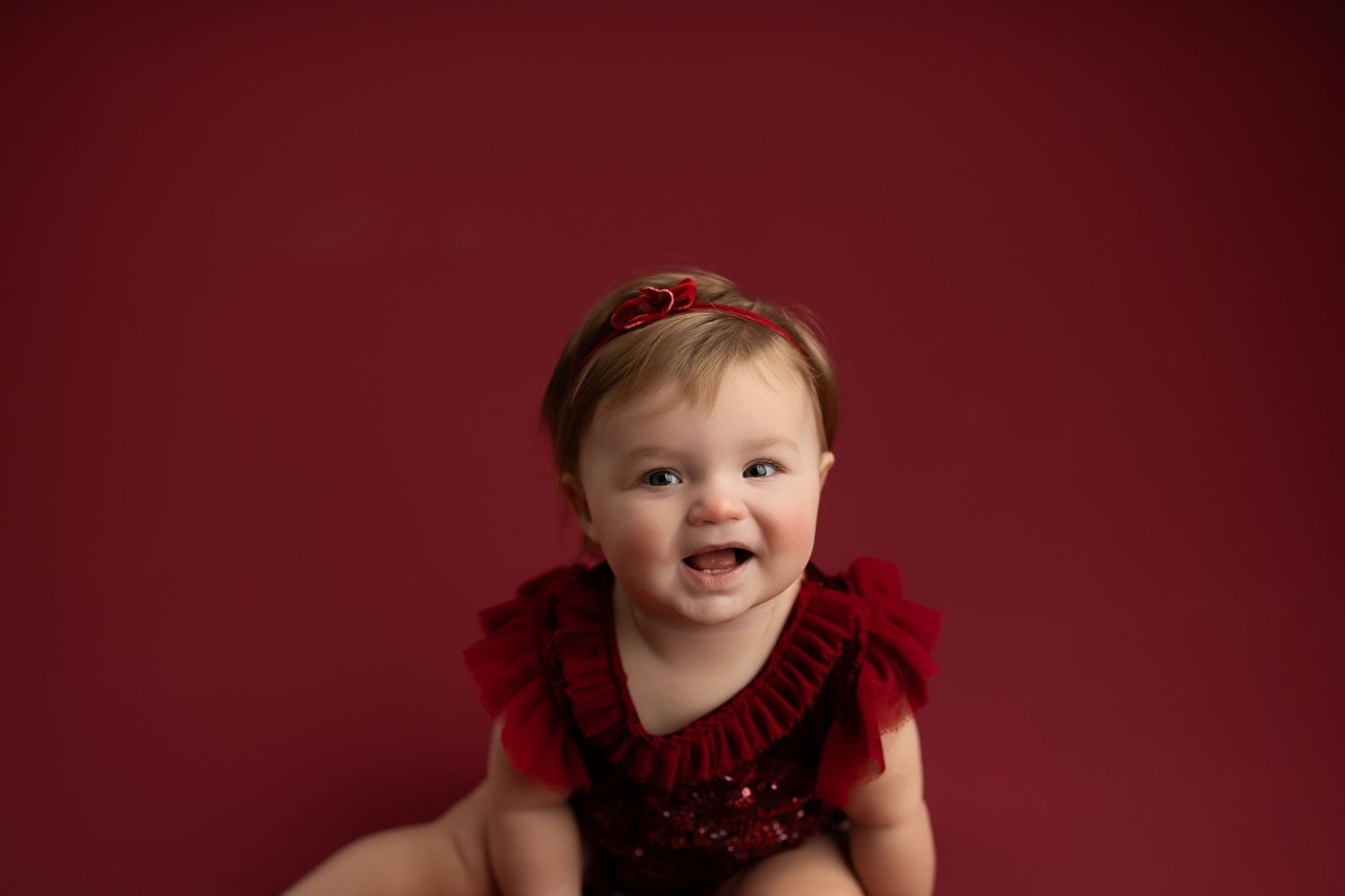 A baby in a red dress laughs joyfully against a deep burgundy background.